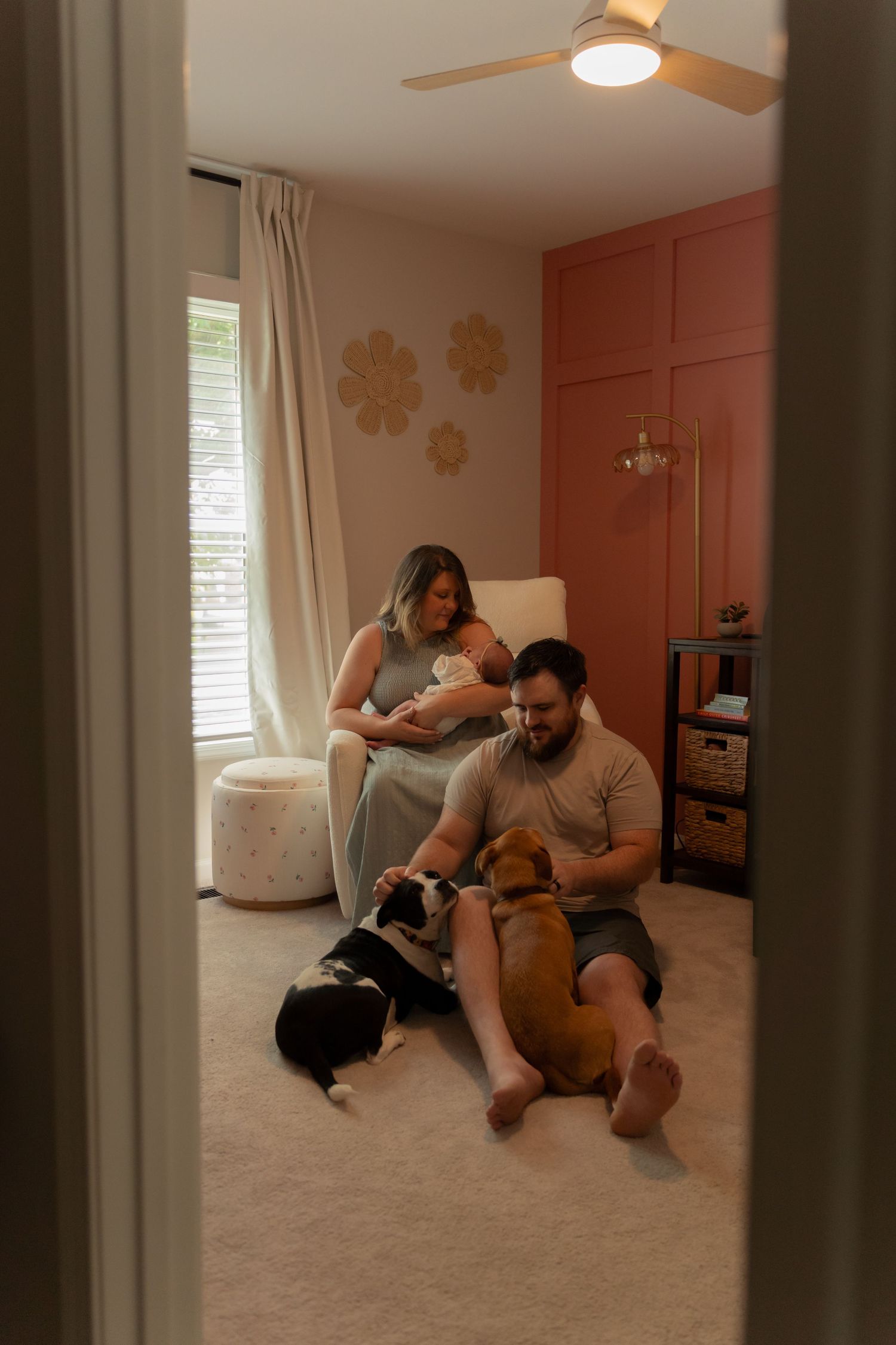 New parents in a pink nursery, mother holding newborn while father sits on floor with two dogs, cozy family moment.