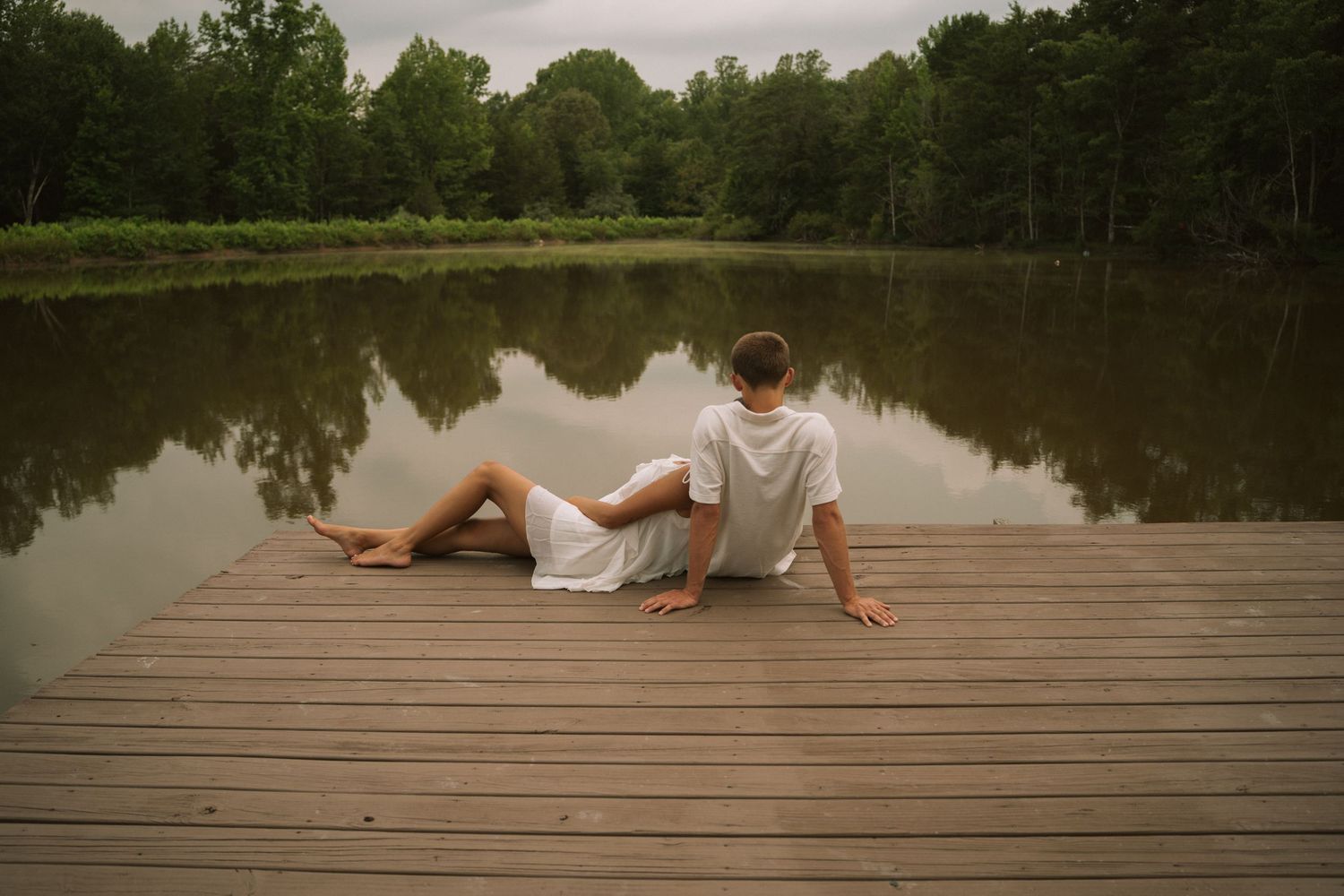 Couple sitting together on a wooden dock overlooking a calm reflective lake surrounded by lush green trees.