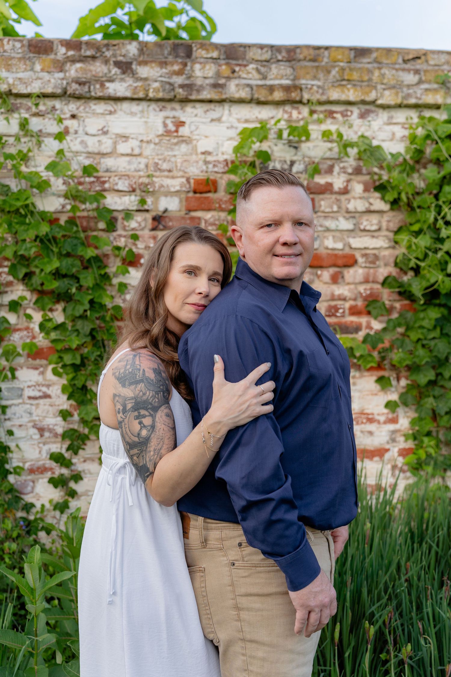 A tattooed woman in a white dress embraces a man in a navy shirt before a rustic brick wall with ivy. Taken by Row And Co Photography
