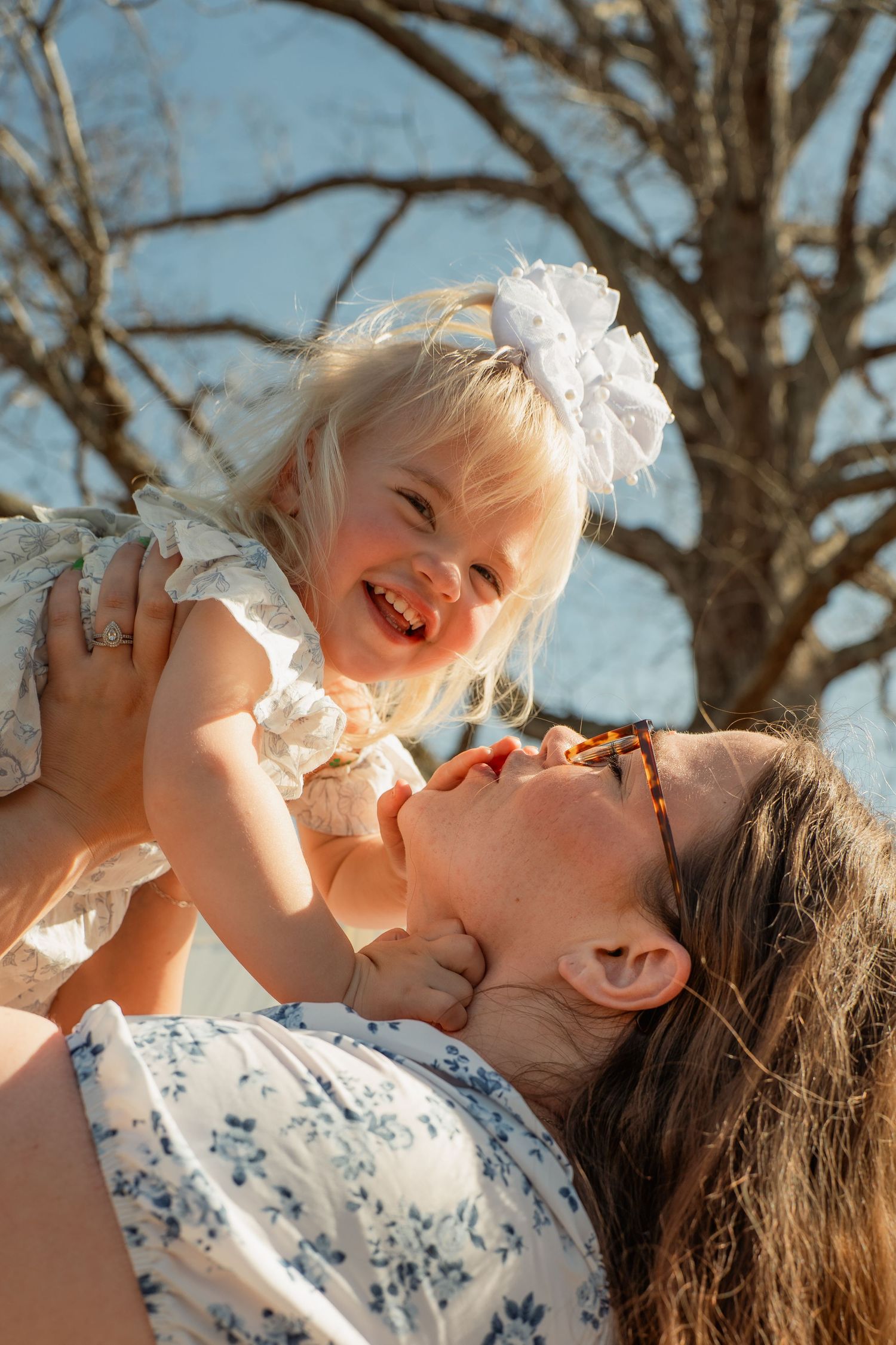 Joyful blonde toddler girl with white bow being lifted by her smiling mother outdoors near bare trees. Taken by Row And Co Photography