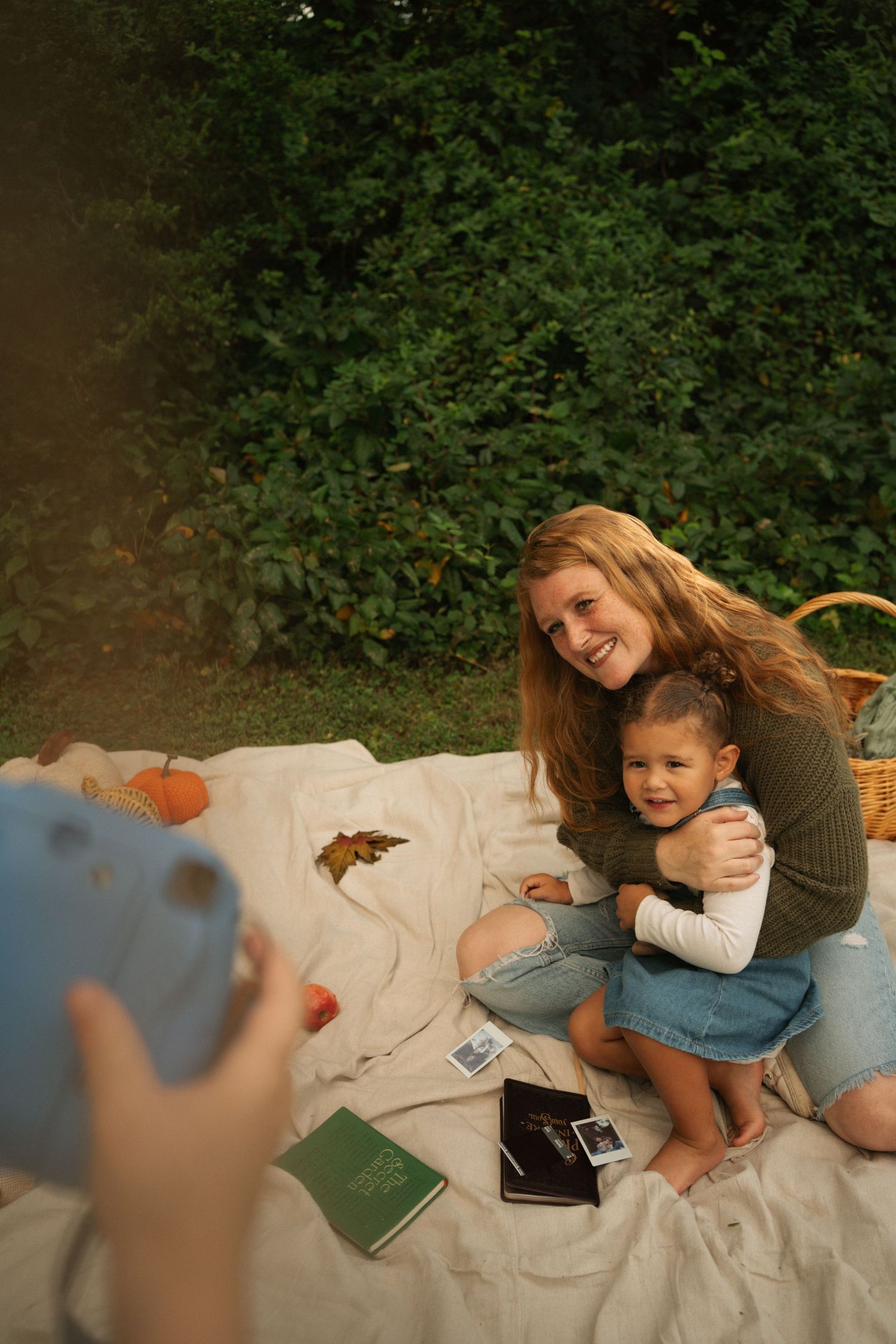 Woman and toddler smile for a polaroid photo on a blanket outdoors during an autumn picnic.