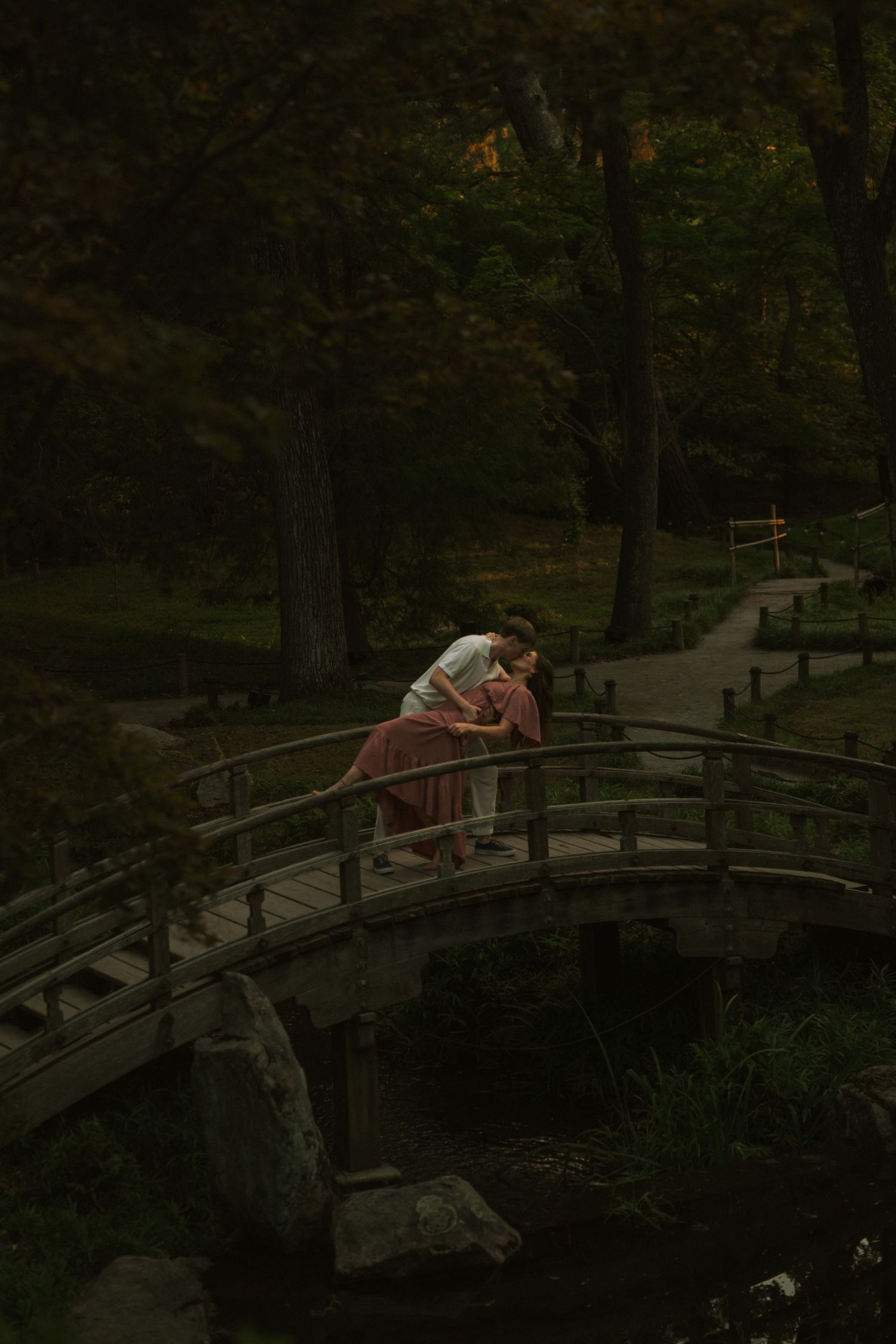A couple embraces on a wooden arched bridge surrounded by lush green trees in a serene park setting.