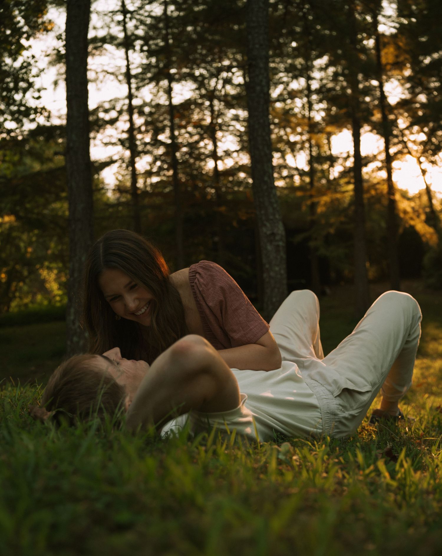 Couple relaxing in a sunlit forest meadow at golden hour, woman smiling down at man lying on grass.