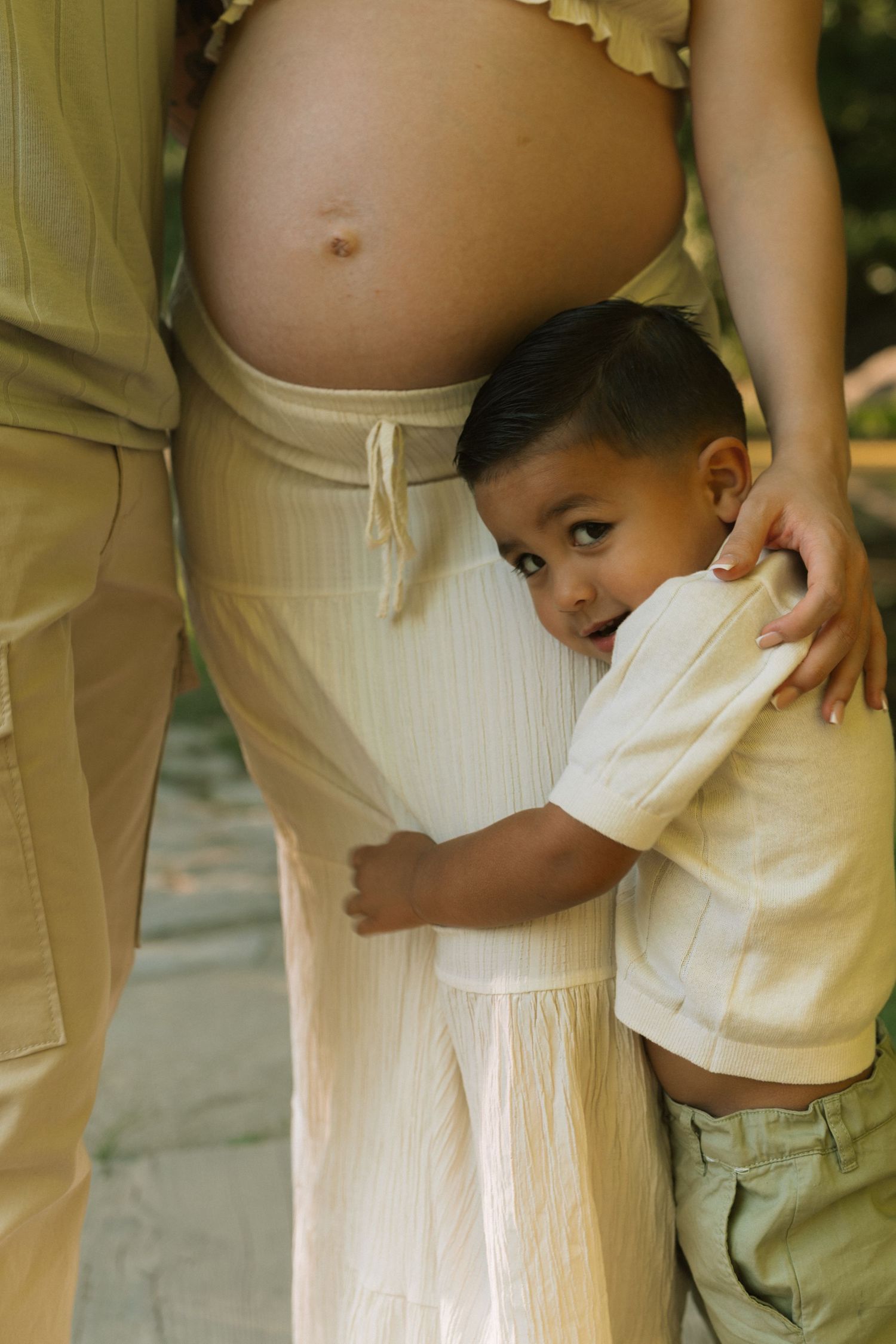 Young boy hugging pregnant mother's belly, wearing cream outfits outdoors.