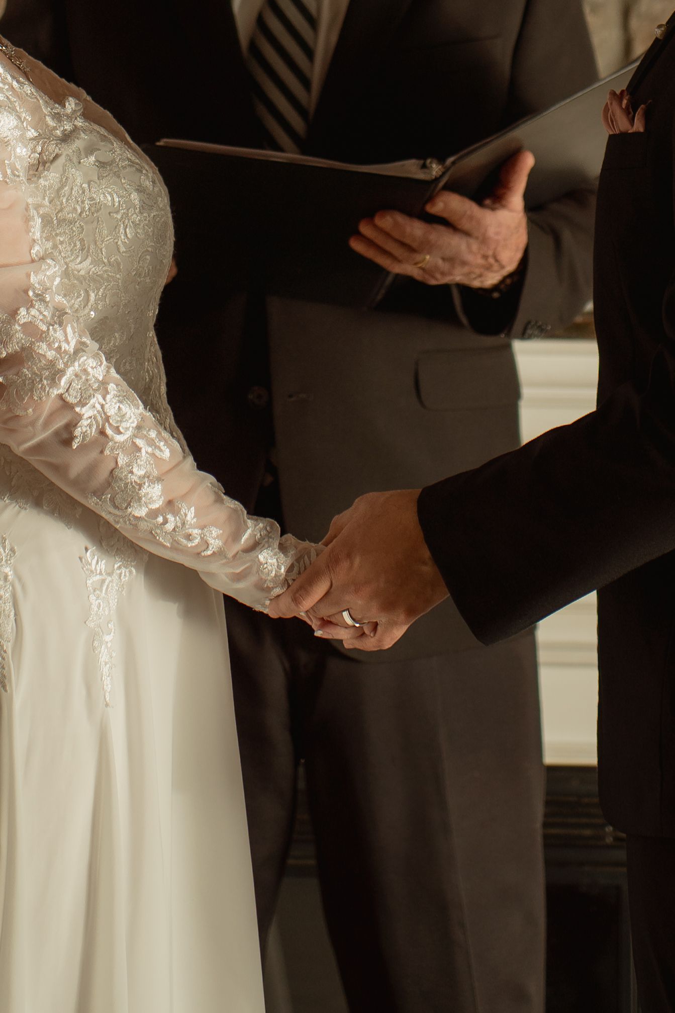 Bride and groom holding hands during wedding ceremony while officiant reads from a book in background.