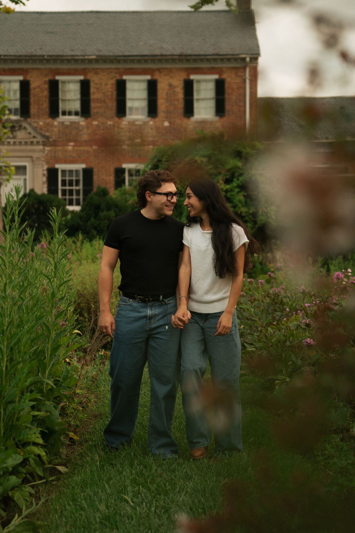 A couple holds hands and smiles at each other in a lush garden in front of a red brick colonial home.