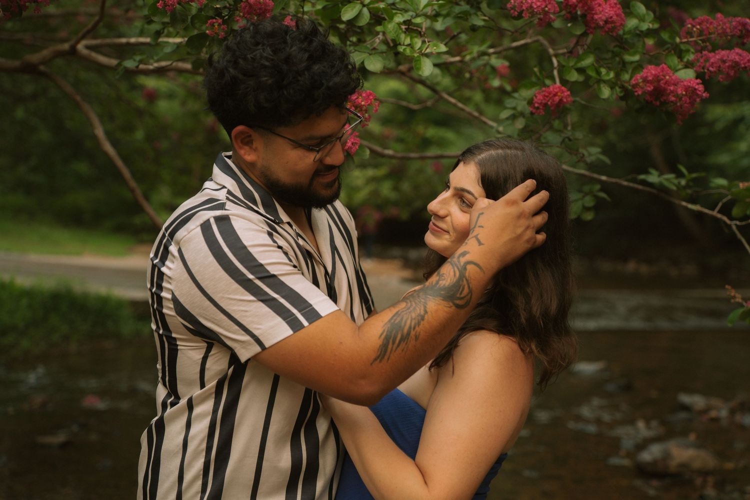 Couple sharing a tender moment under pink blooming tree by a creek, man gently holding woman's face. Row and Co Photography