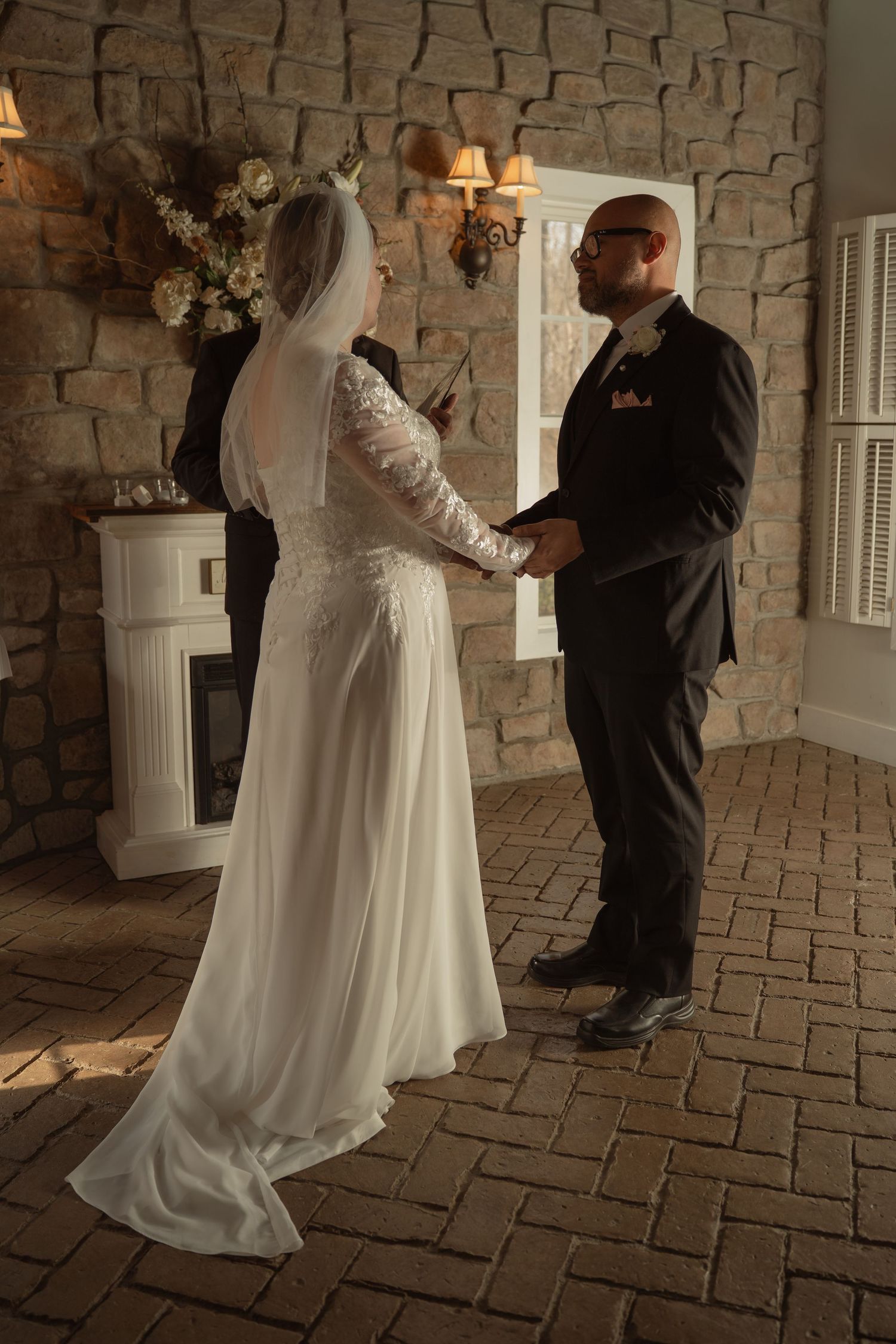 Bride and groom exchange vows indoors at rustic stone wall venue, bride in lace gown with veil, groom in dark suit.