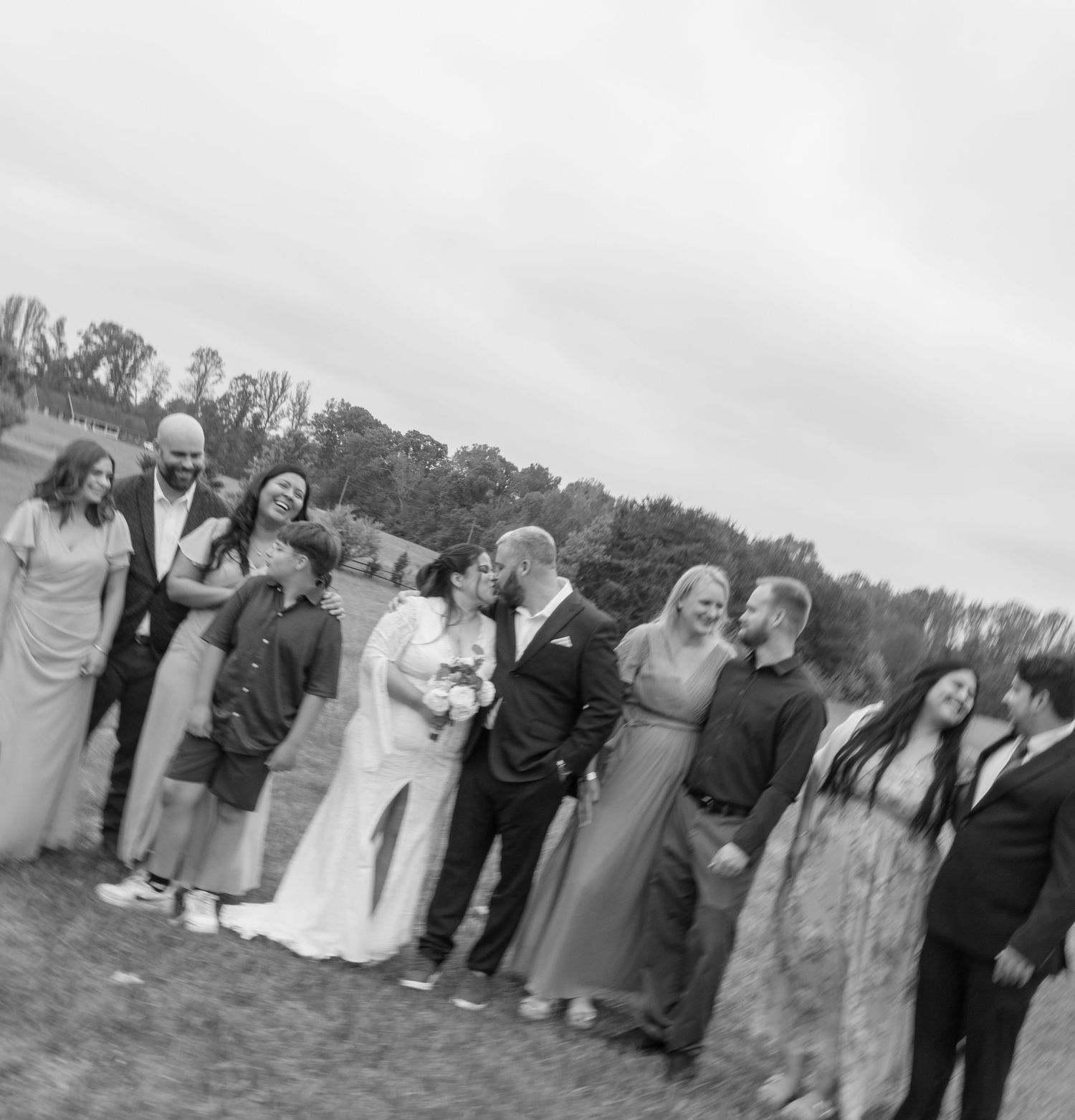 Black and white wedding photo of bride and groom kissing surrounded by family and wedding party on grassy outdoor venue.