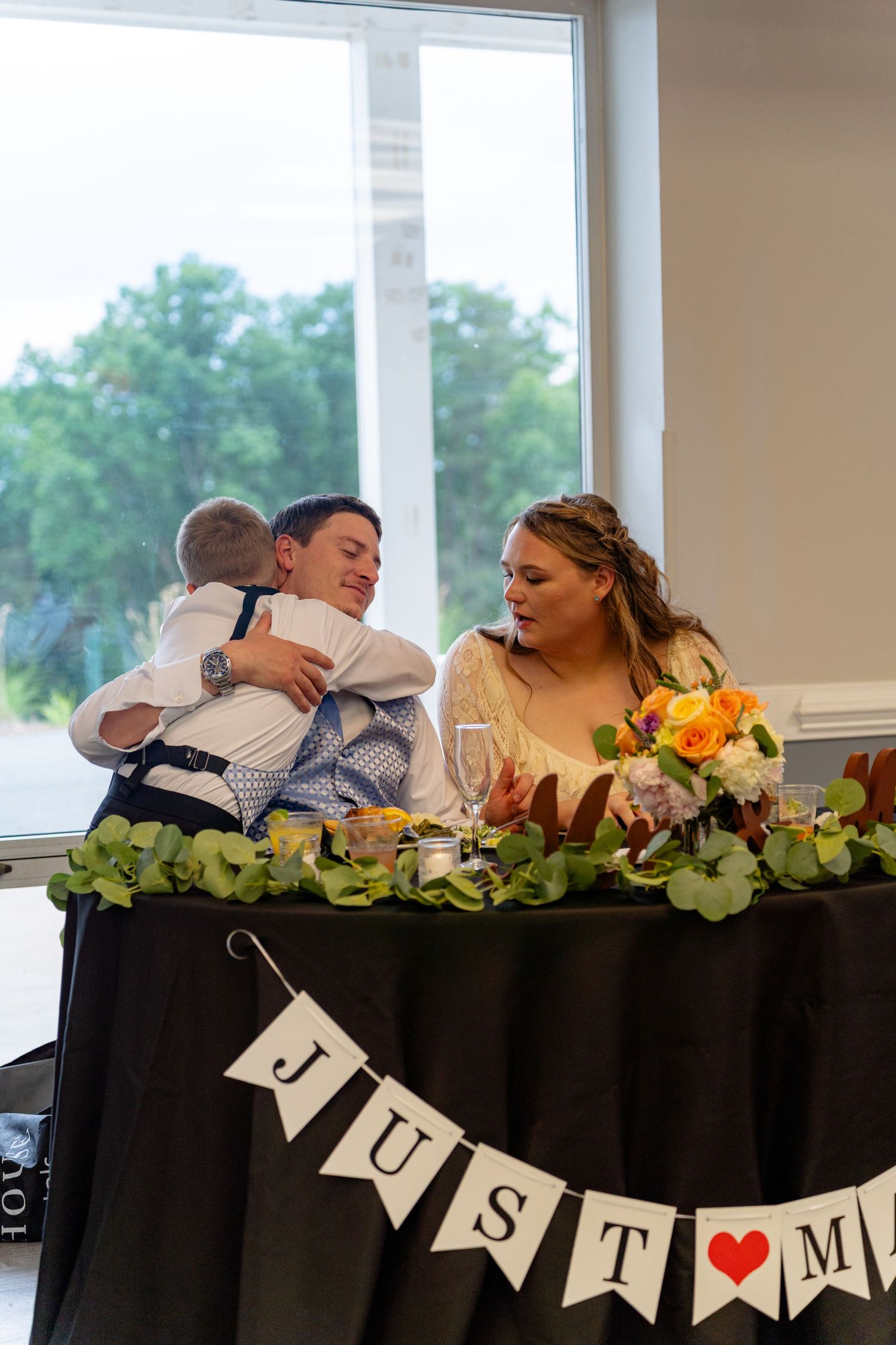 Newlyweds at wedding reception table with Just Married banner, bride smiles as groom hugs child among floral decorations.
