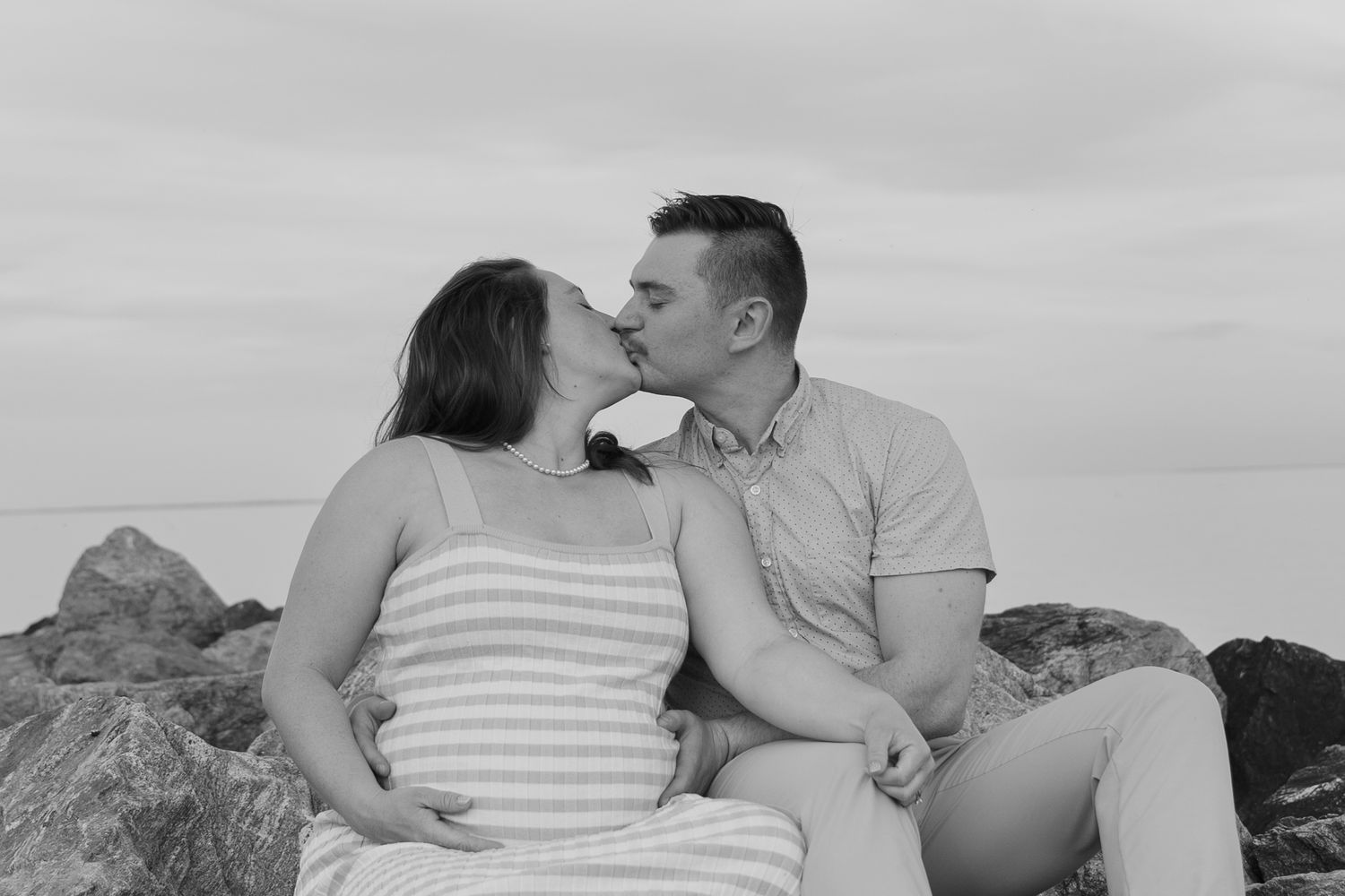 Black and white photo of couple kissing romantically while sitting on rocks by the ocean. Taken by Row And Co Photography in FXBG VA