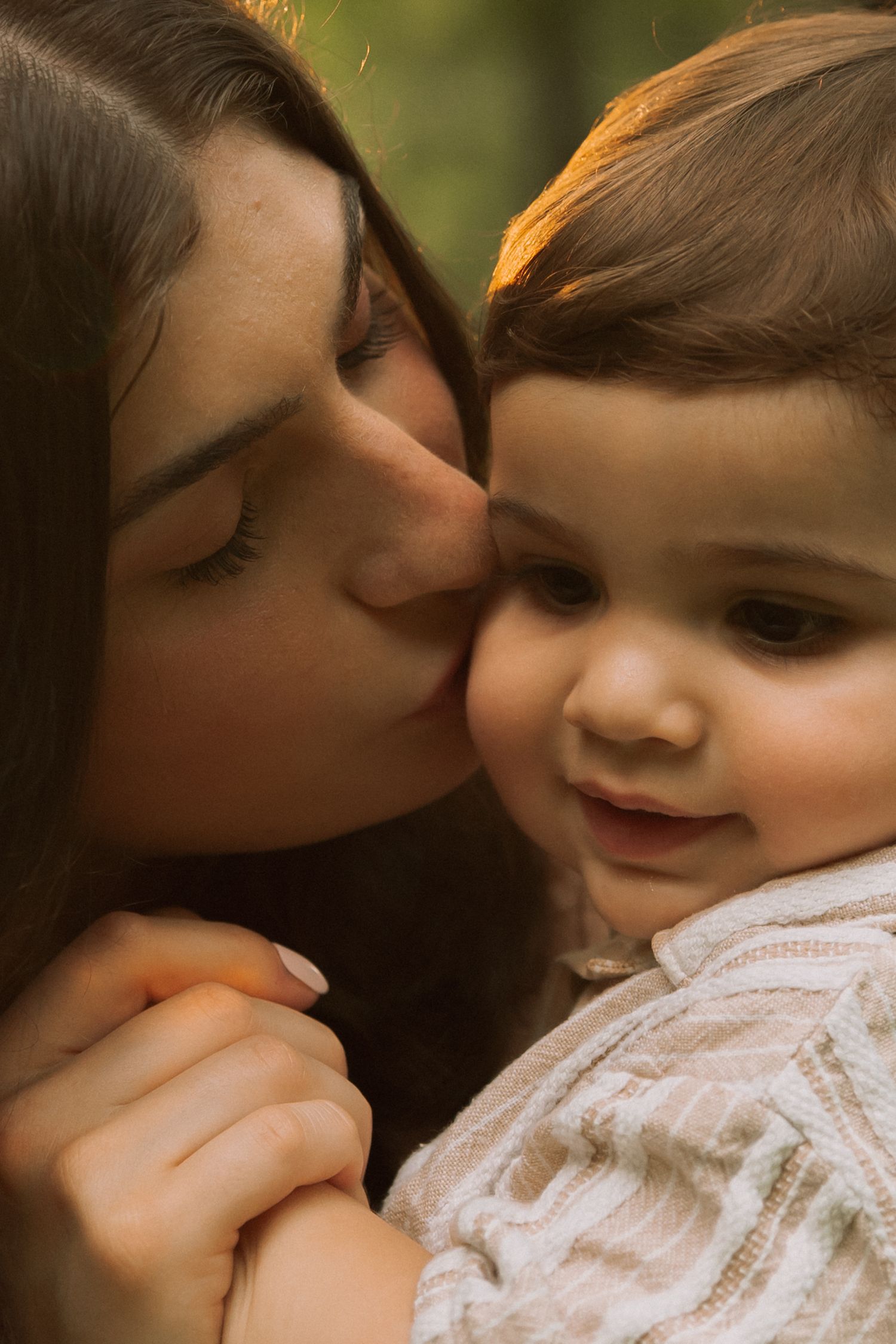 A mother tenderly kisses her baby's cheek outdoors in warm golden light. Row and Co Photography