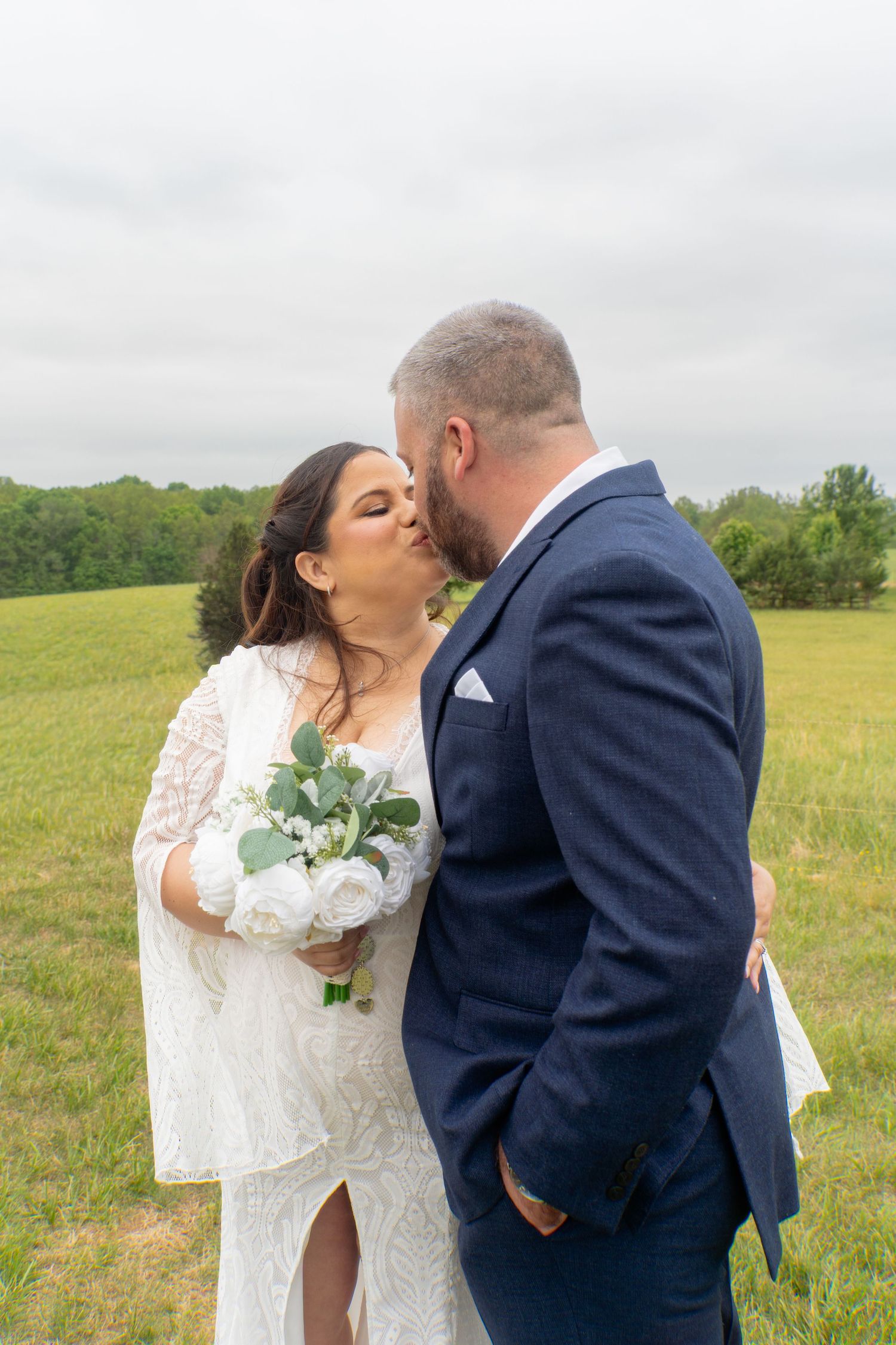 Bride and groom sharing a kiss in a green field, bride holding white floral bouquet in lace wedding dress.