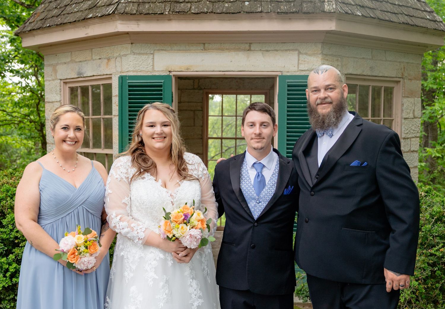 Wedding party of four posing outdoors near a stone gazebo with green shutters, bride holding colorful bouquet.