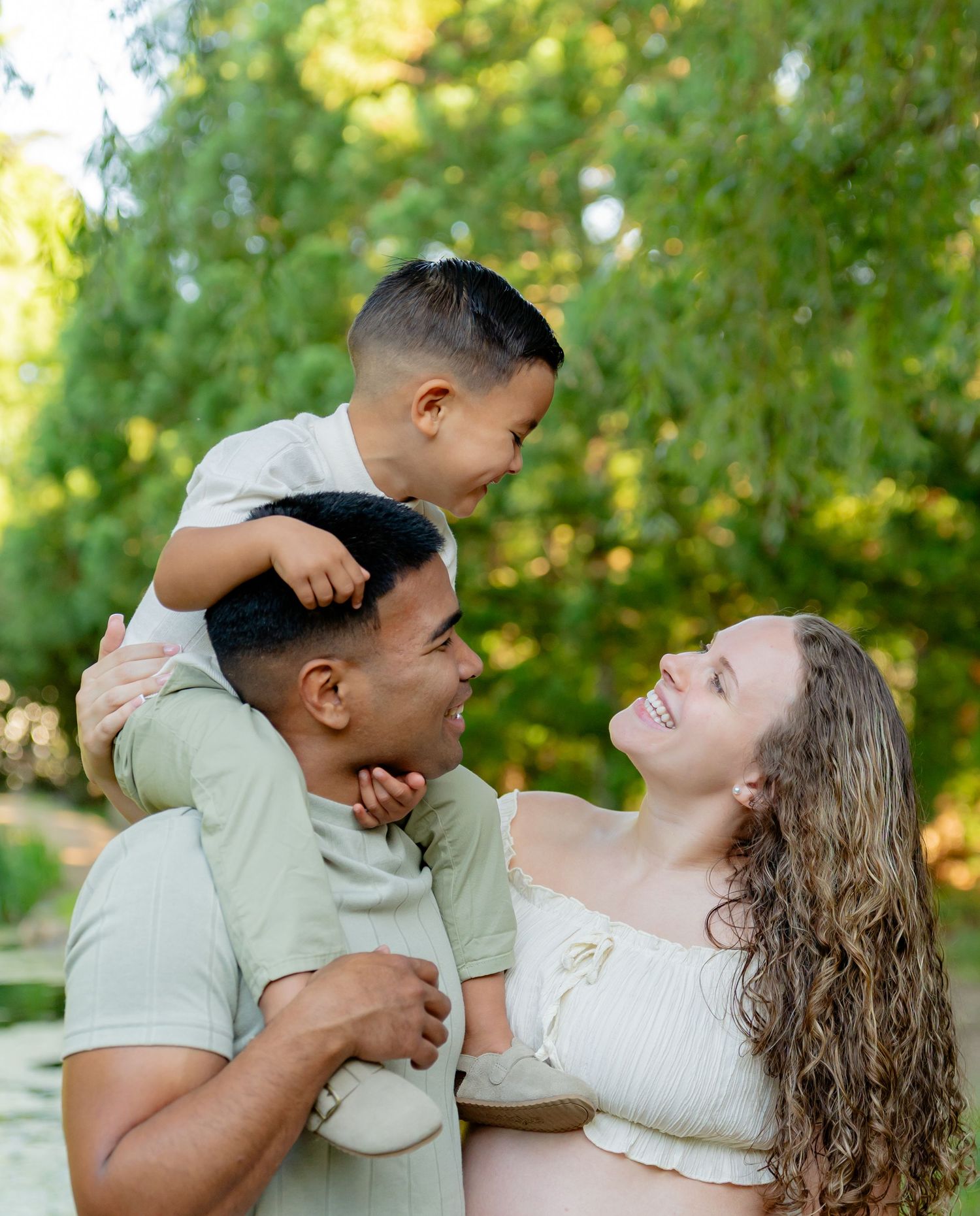 A pregnant woman and her partner smile joyfully as their young son sits on dad's shoulders outdoors. Taken by Row And Co Photography in FXBG