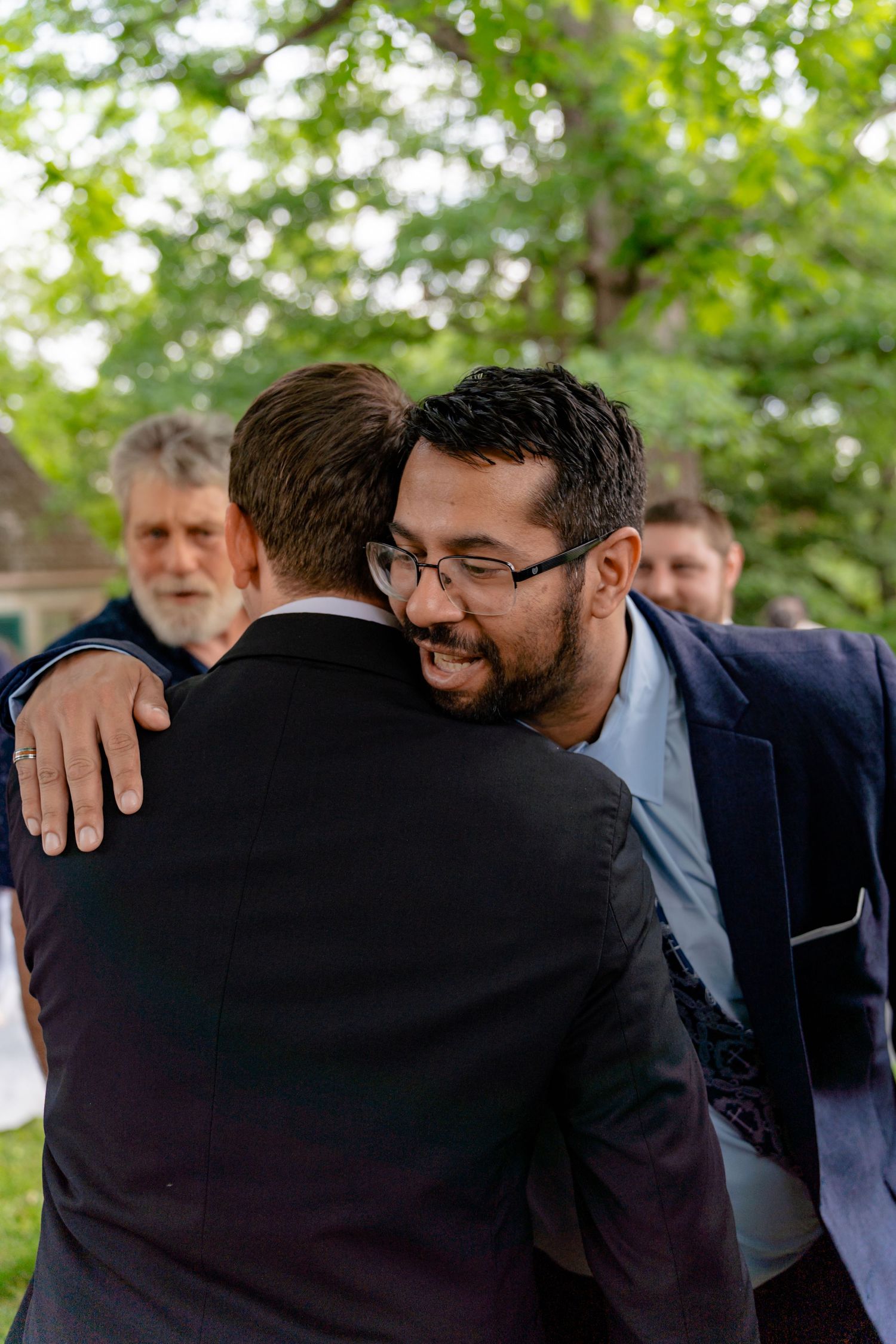 Two men share a warm embrace outdoors at a wedding celebration surrounded by lush green trees.