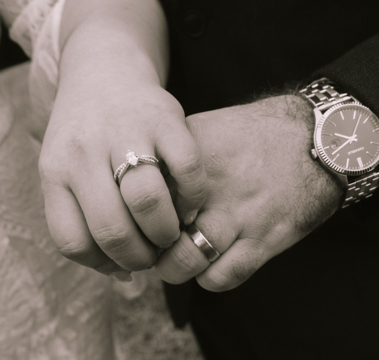 Black and white photo of a couple holding hands, showing an engagement ring, wedding band, and a wristwatch.