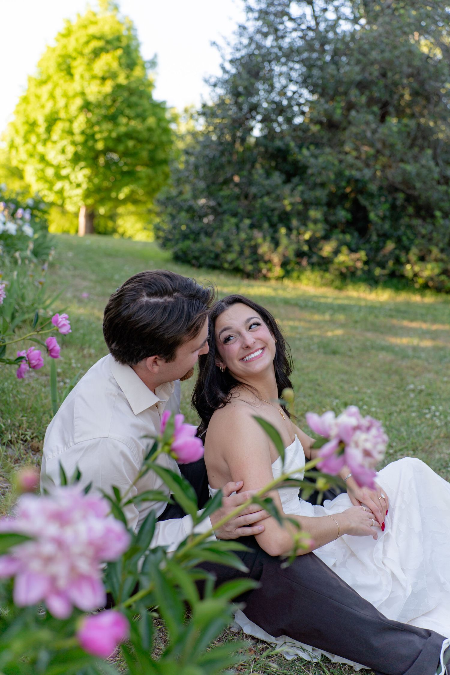 Joyful bride and groom sitting on grass surrounded by pink flowers during sunny outdoor wedding. Taken by Row And Co Photography in FXBG VA