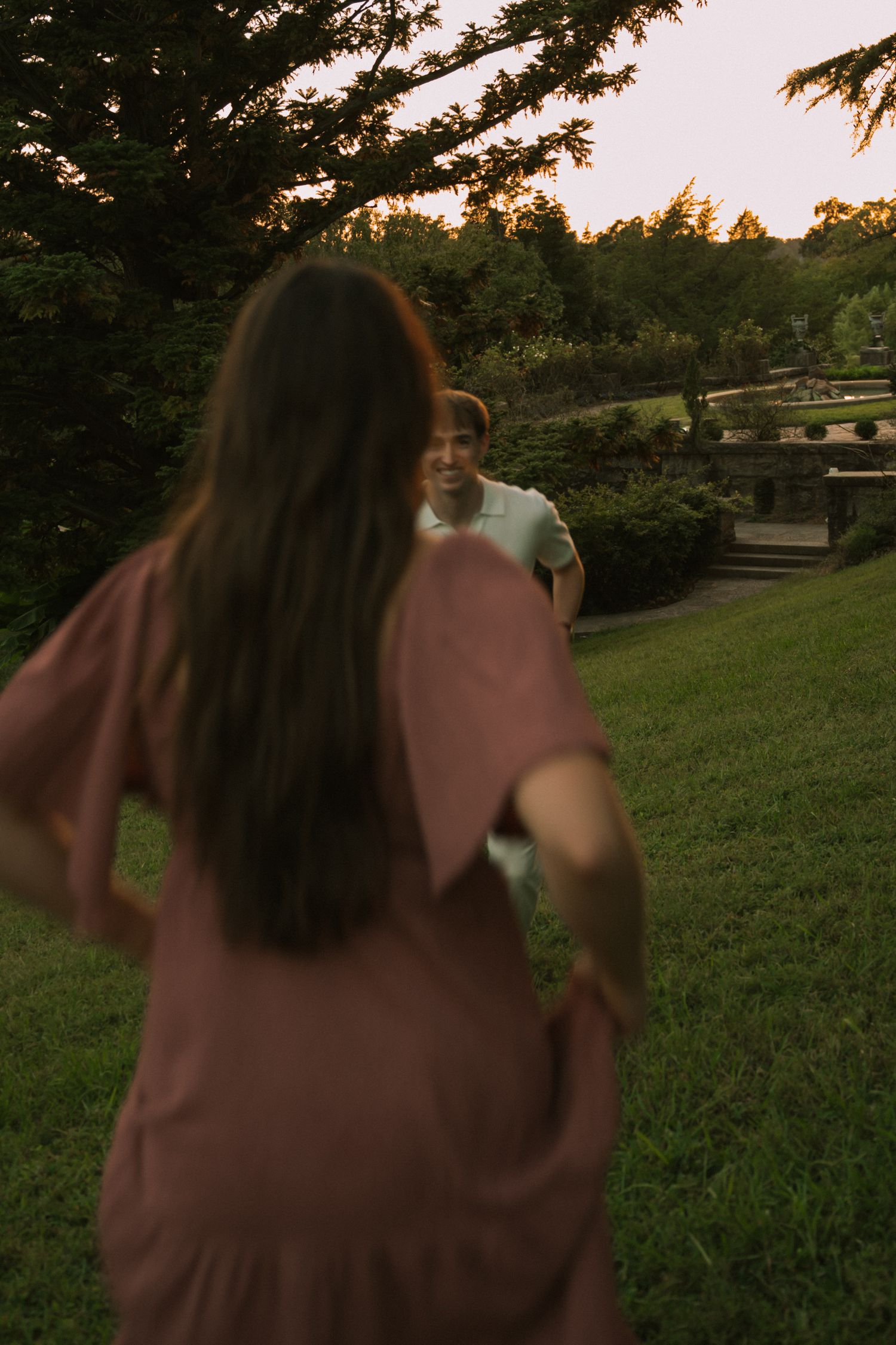 A woman in a pink dress reaches toward a smiling man in a lush green park at golden hour sunset.