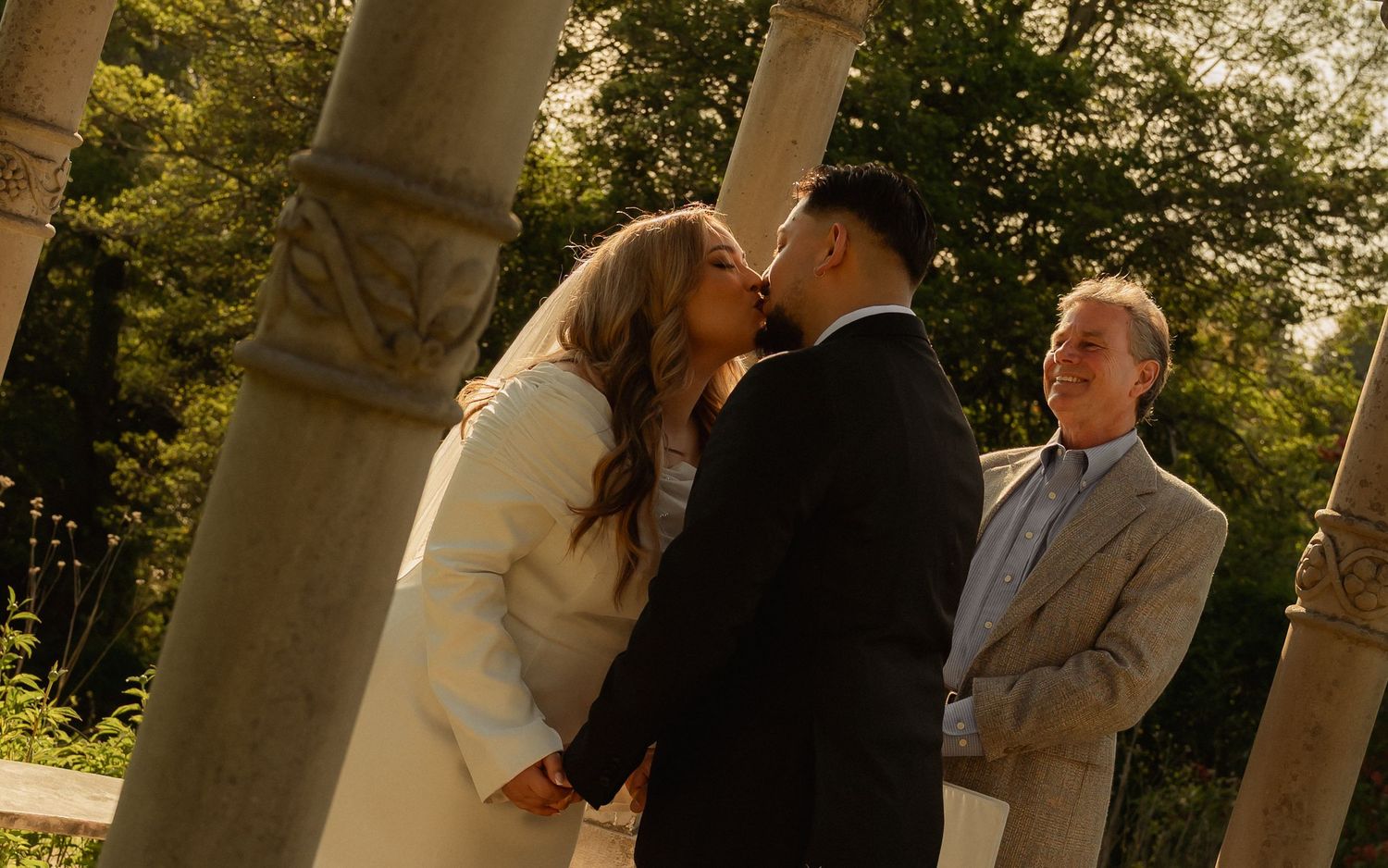 Couple sharing a romantic kiss during outdoor wedding ceremony with officiant and stone columns in lush garden setting.