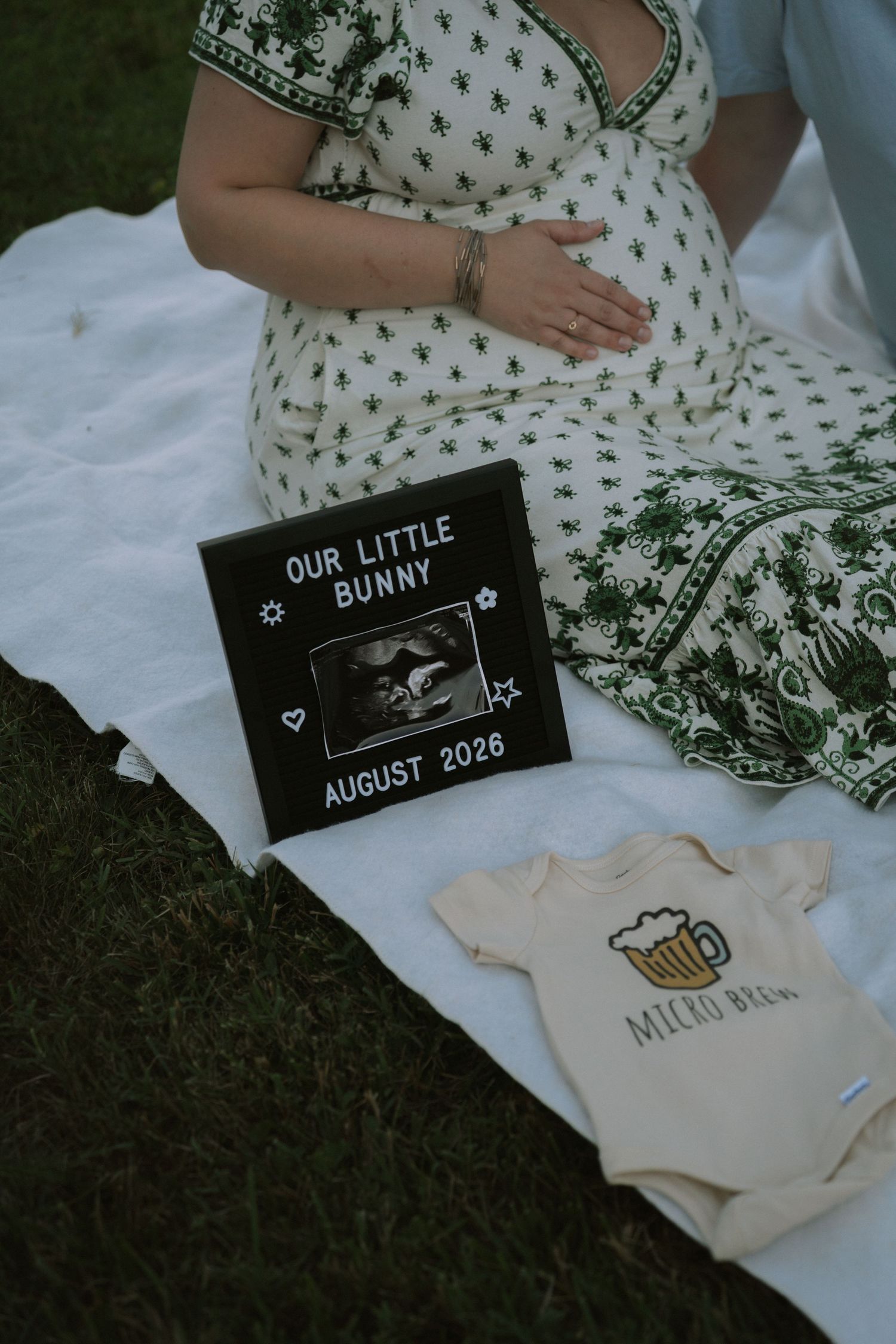 Pregnancy announcement with ultrasound photo, 'Our Little Bunny August 2026' sign, and baby onesie on white blanket.
