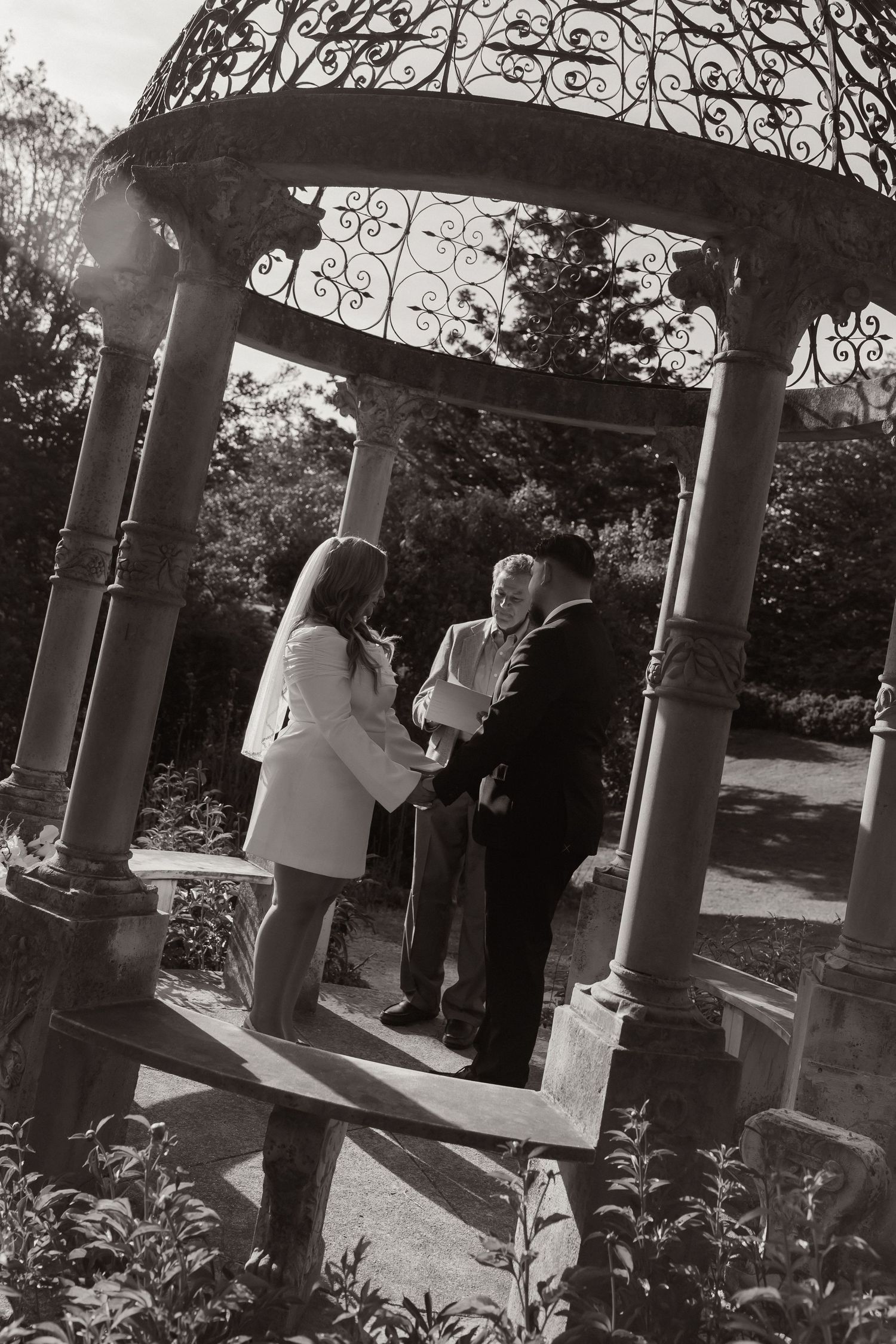 Black and white photo of a couple exchanging vows under an ornate iron gazebo in a lush garden setting.