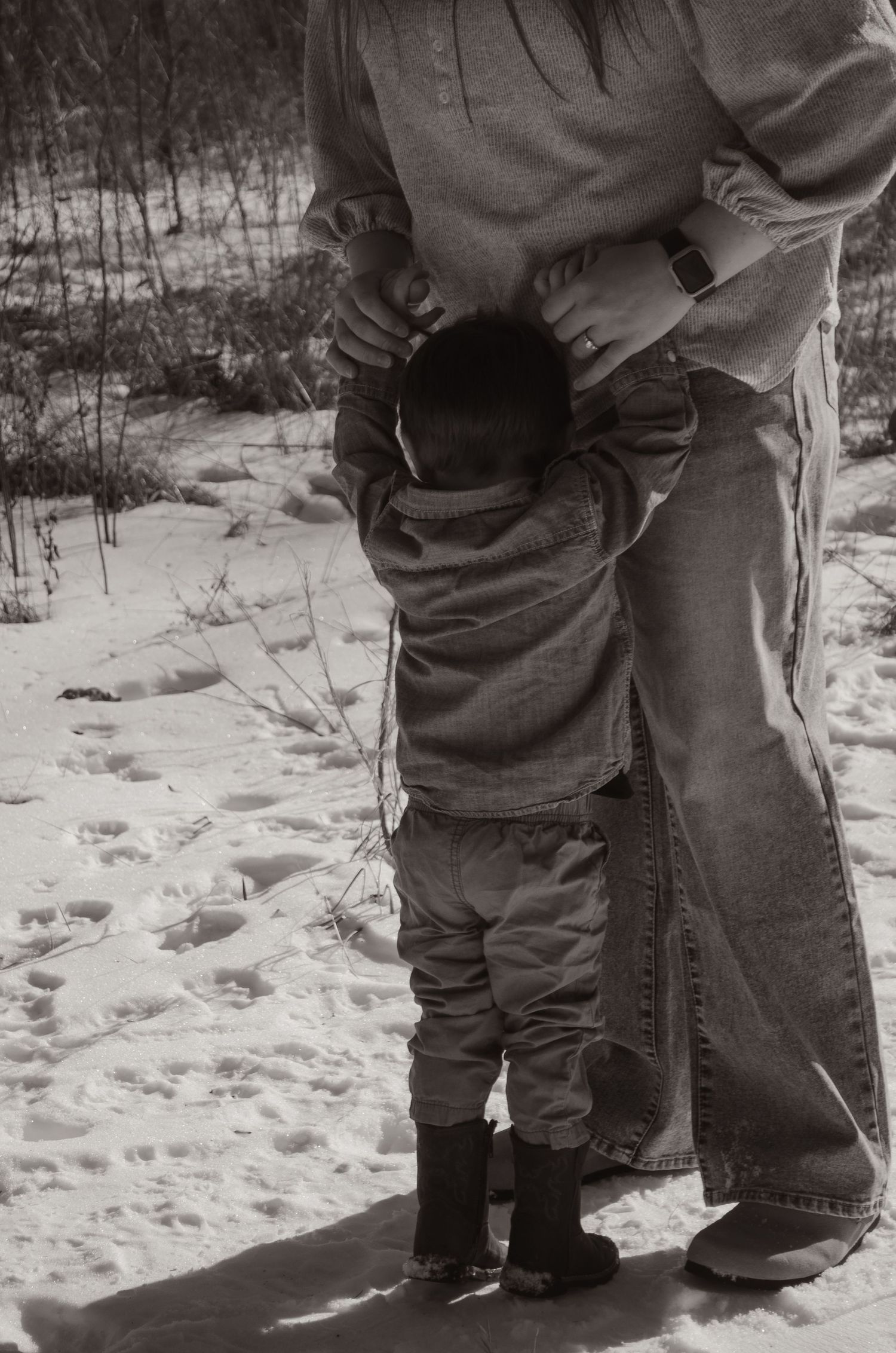 Black and white photo of a toddler clinging to a parents legs in a snowy field. Taken by Row And Co Photography in FXBG VA