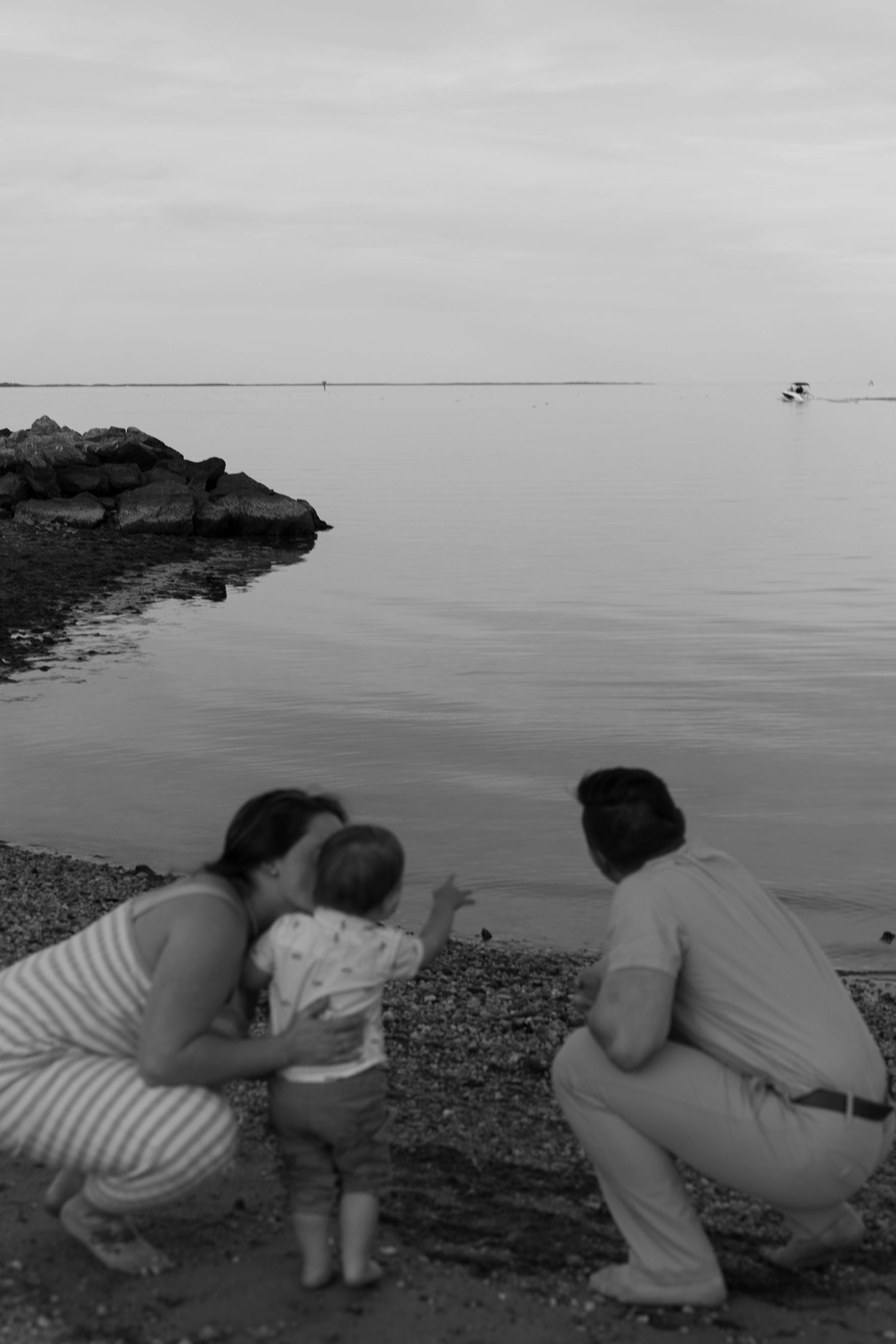 Black and white photo of a family with a baby crouching by the calm water at the beach. Taken by Row And Co Photography in FXBG VA