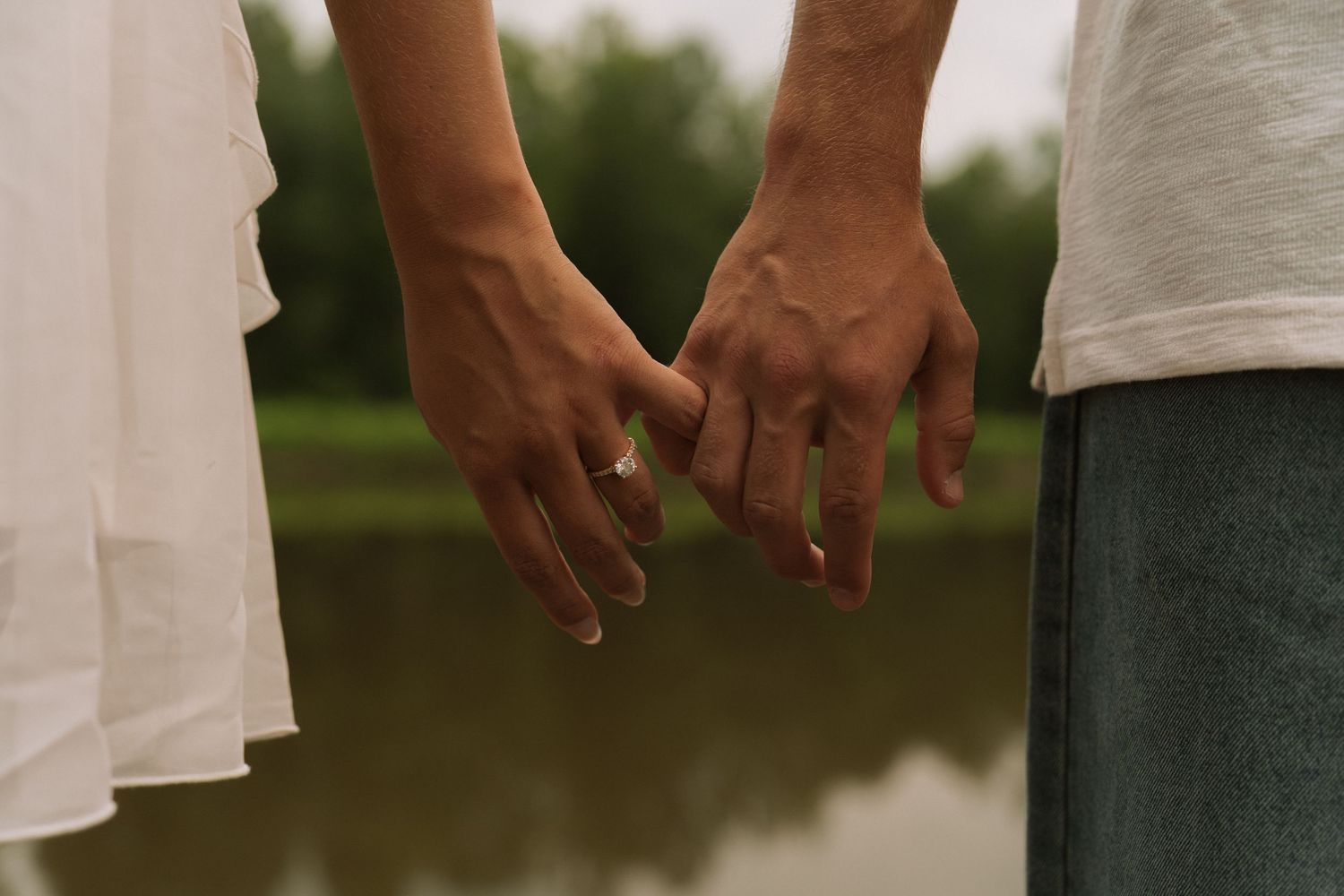 Couple holding hands with intertwined fingers near a lake, wearing white shirts, with an engagement ring visible.