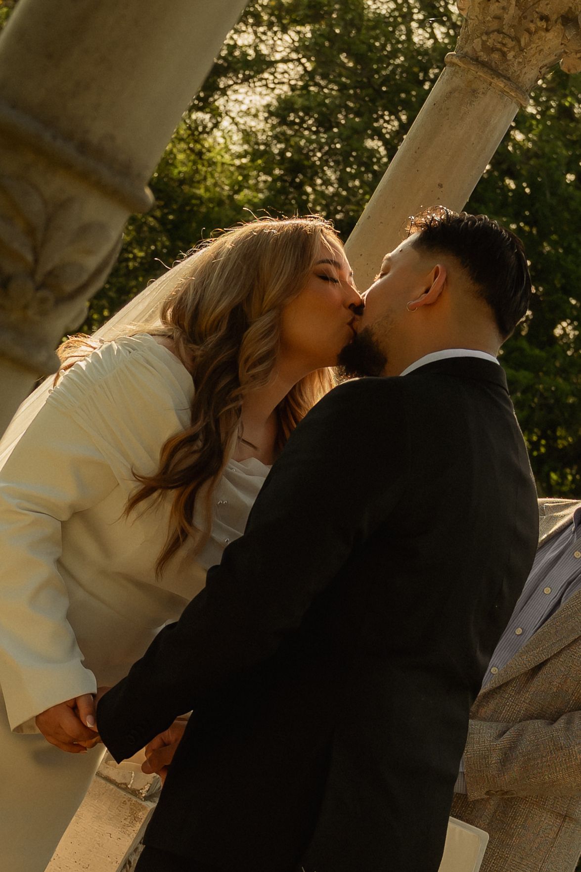Bride and groom sharing a romantic kiss outdoors, surrounded by stone columns and lush green trees in golden hour light.