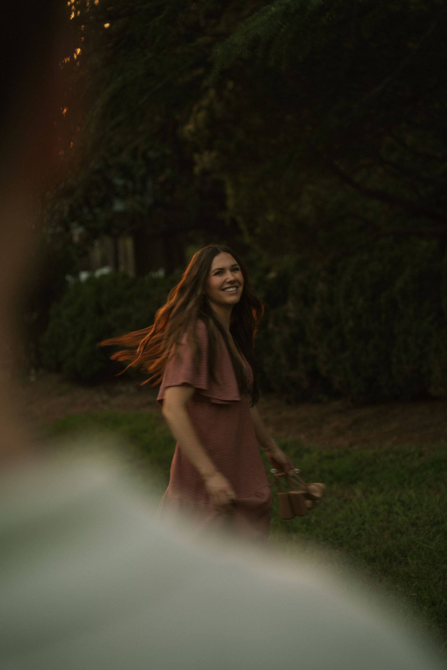 Smiling woman in pink dress with flowing hair walks outdoors near lush green trees at dusk.