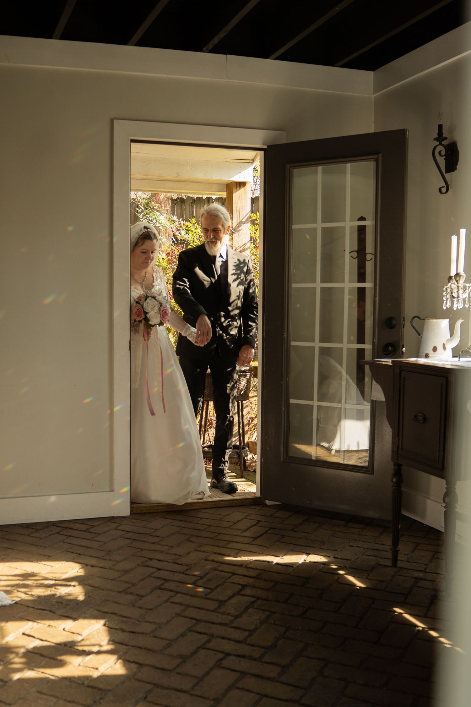 Bride in white gown and groom in dark suit entering through doorway into sunlit brick-floored room on wedding day.