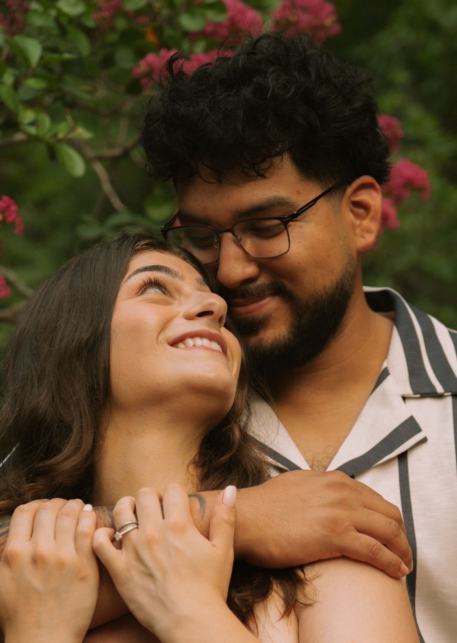 A smiling couple embraces outdoors near pink flowering bushes, woman wearing a ring looks up lovingly at bearded man with glasses. Row and C