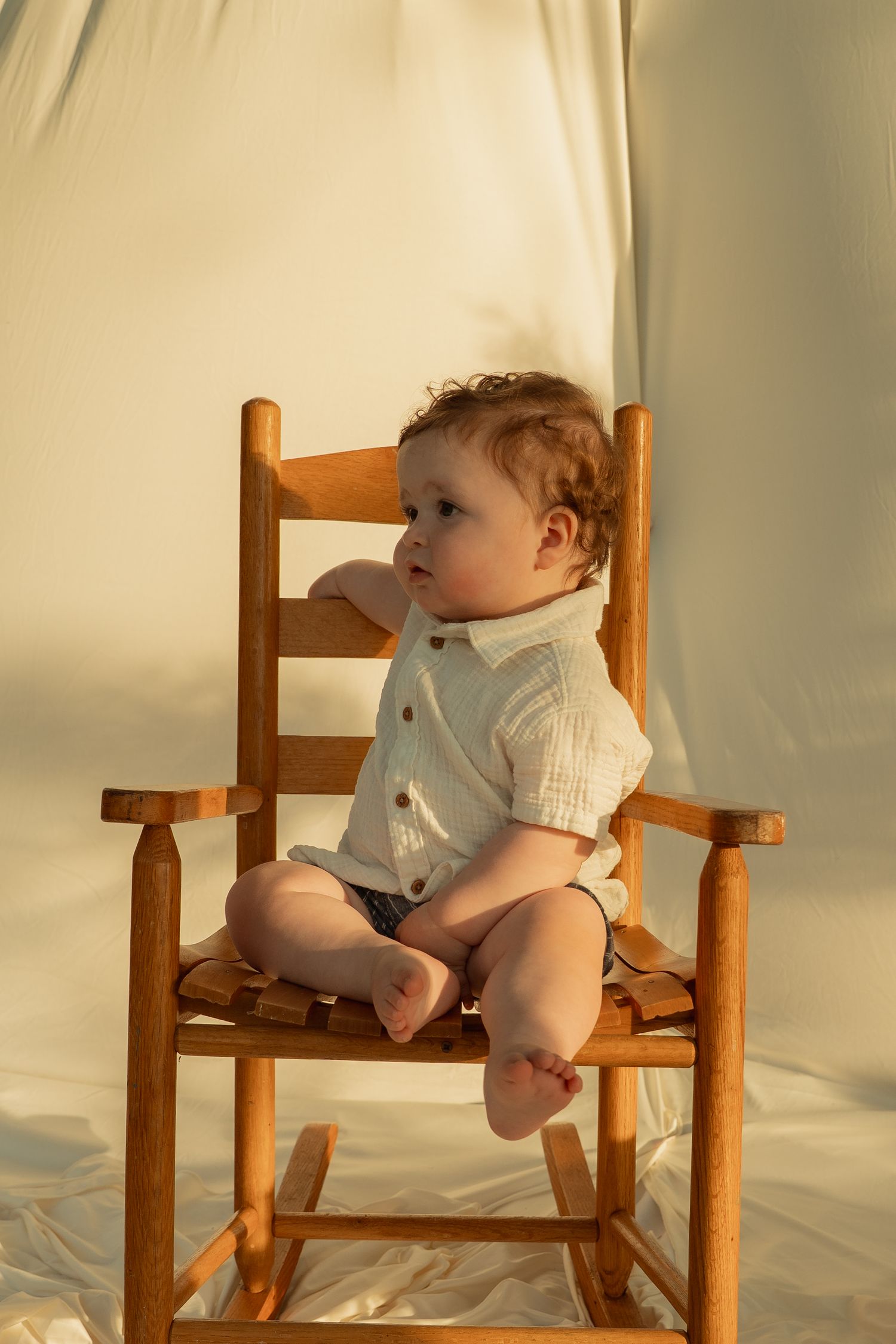 Adorable baby in white button shirt sitting in small wooden rocking chair in warm golden light. Taken by Row And Co Photography in FXBG VA