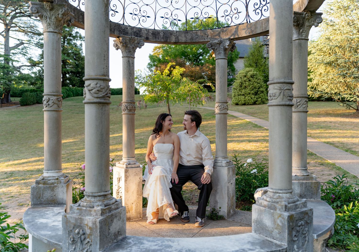 Couple sitting together under an elegant stone gazebo surrounded by lush gardens at a romantic outdoor wedding venue.