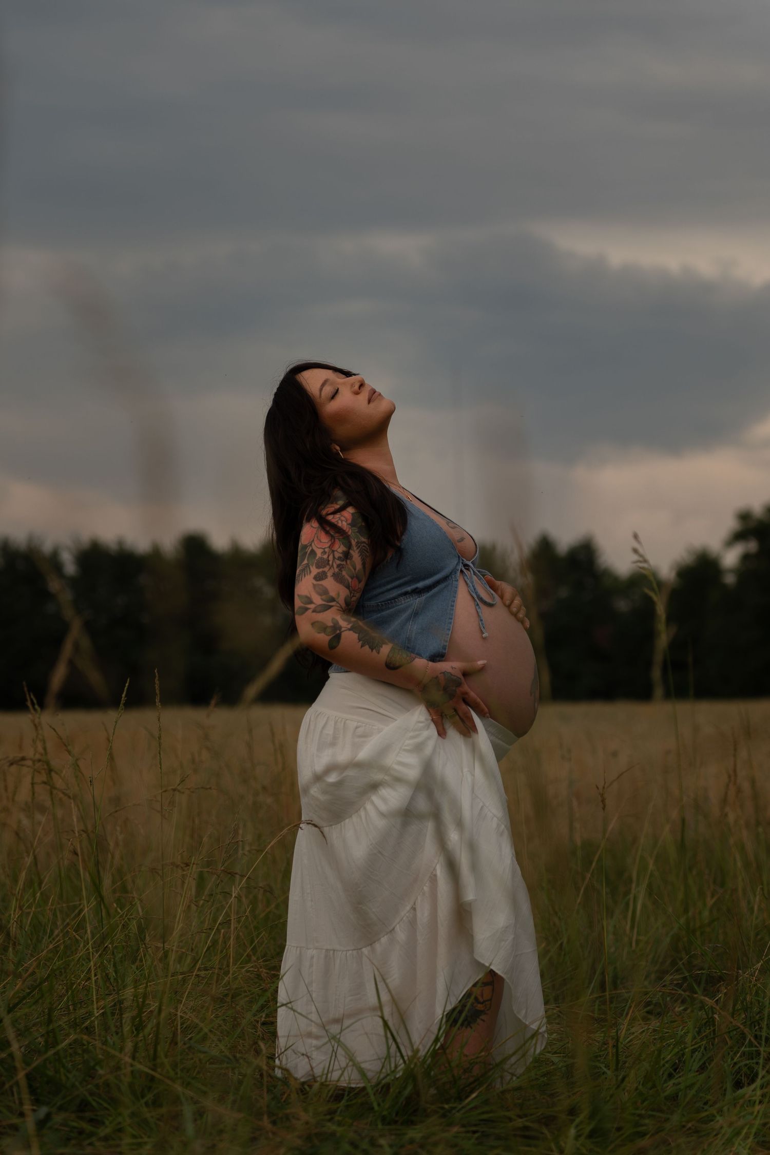 Pregnant woman with tattoos in white skirt and blue top standing in field, cradling baby bump under cloudy sky.