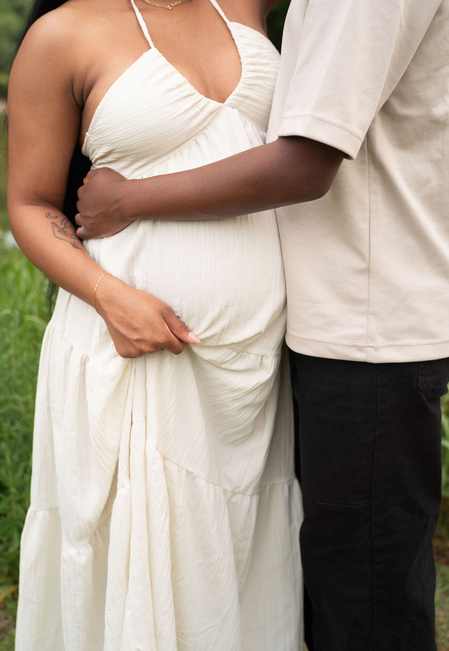 A pregnant woman in a white halter maxi dress stands with her partner's arm around her waist outdoors.