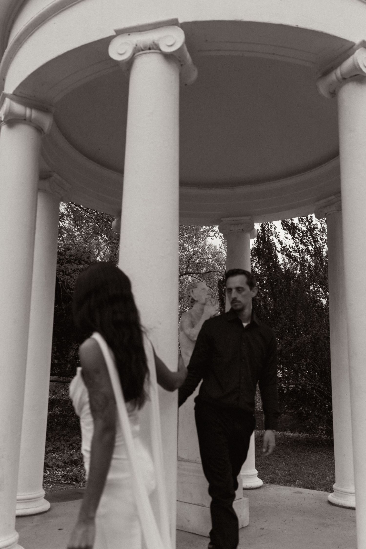 Black and white photo of a couple holding hands around a white column in an elegant outdoor gazebo. Taken by Row And Co Photography in FXBG