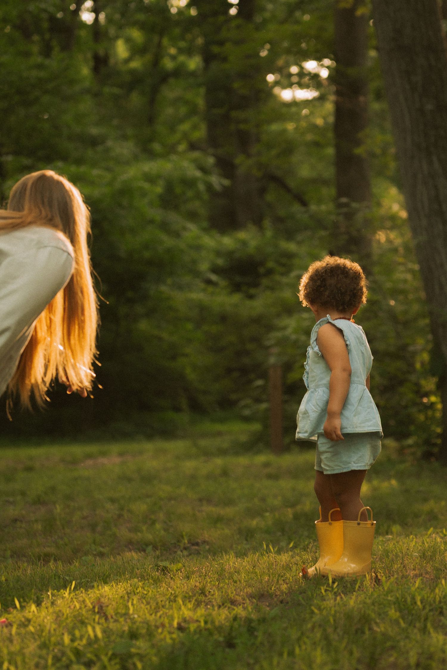 Young child in yellow rain boots stands on grass watching another child play in golden sunset light. Taken by Row And Co Photography in FXBG