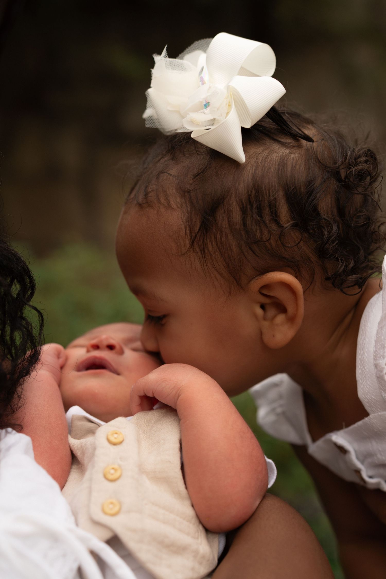 A young toddler girl with a white bow kisses a newborn baby held in someone's arms outdoors.