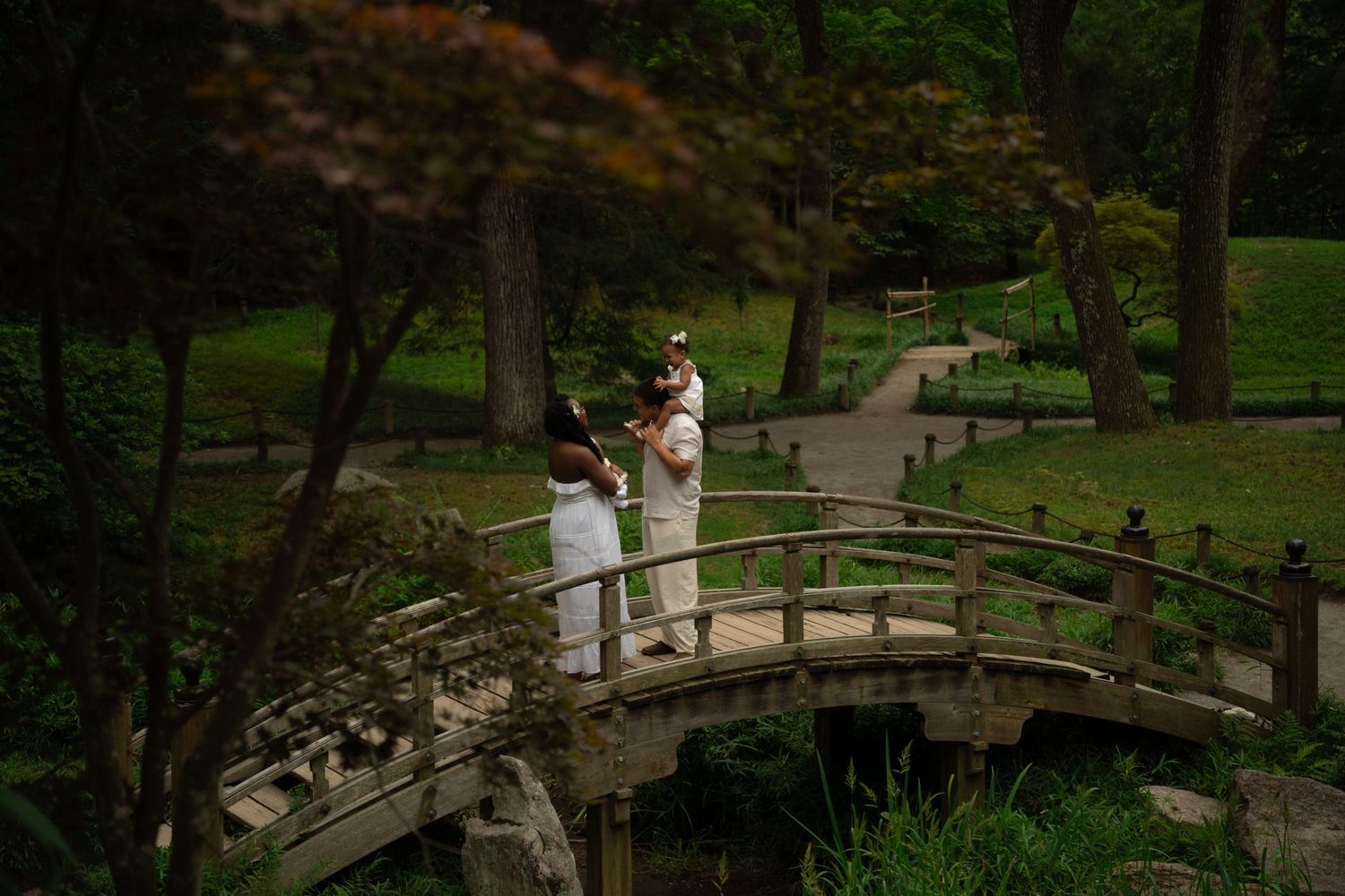 Couple in white attire standing on a wooden arched bridge in a lush Japanese garden.
