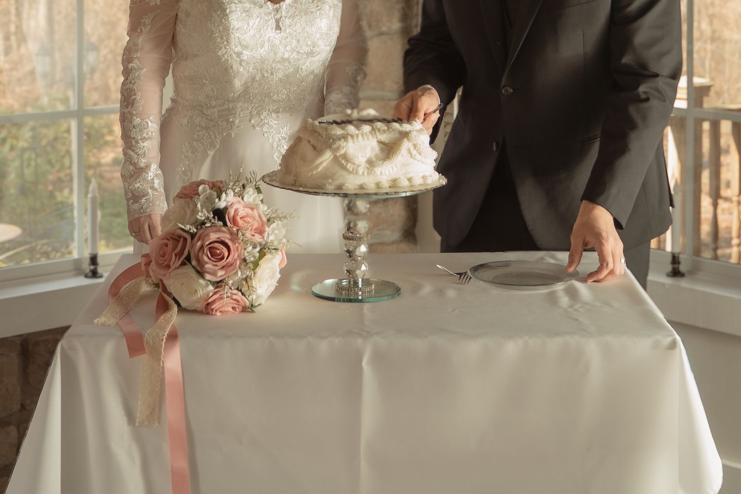 Bride and groom cutting a white wedding cake on a table with pink roses bouquet and satin ribbon.
