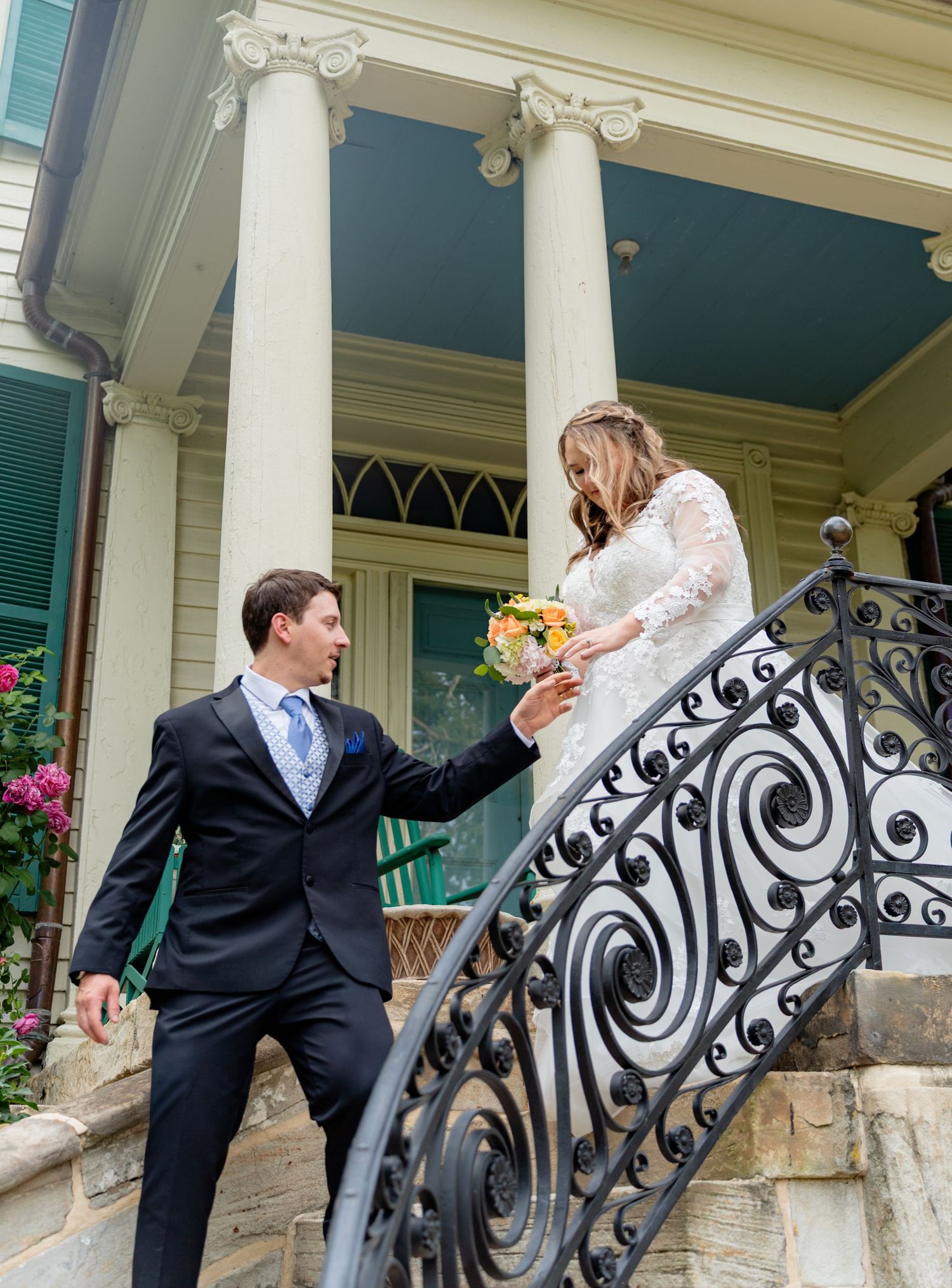 Bride and groom on ornate iron staircase of elegant Victorian home, groom holding bride's hand as she descends.