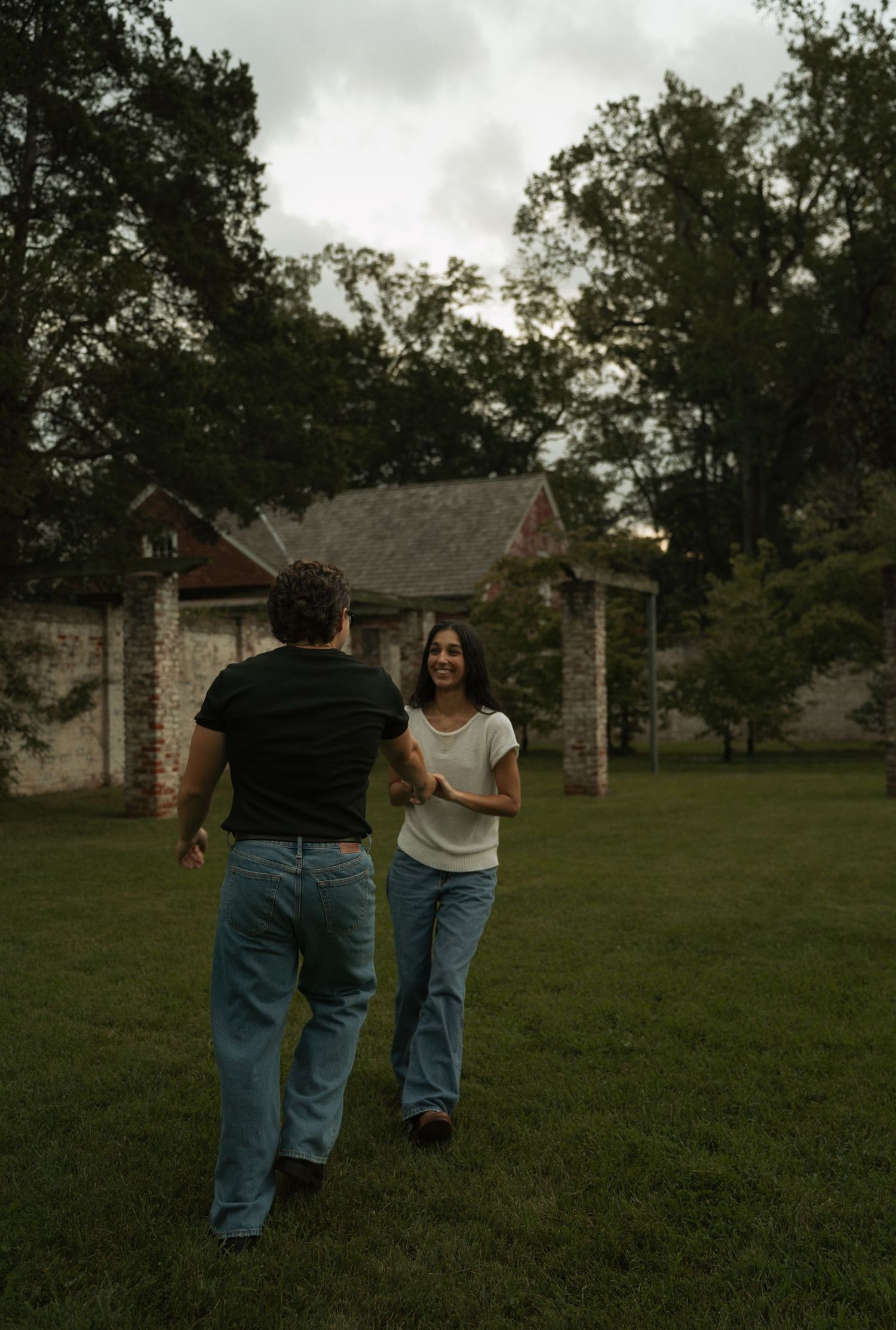A couple walks toward each other on a grassy lawn at dusk, smiling, with trees and a brick building in the background.