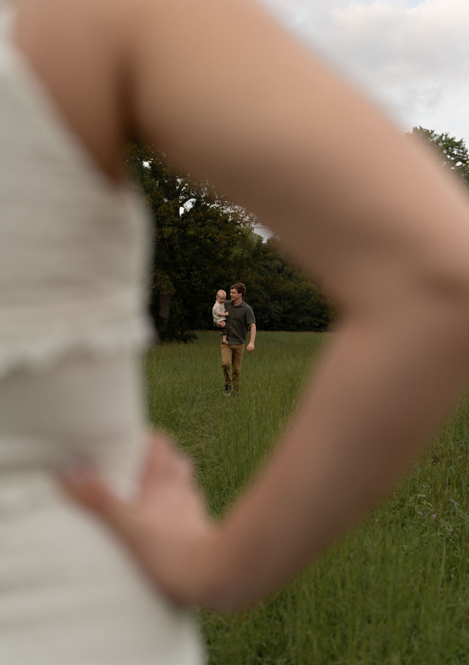 Bride in white dress framing a man holding a child in a lush green field, captured in a romantic outdoor wedding photo.