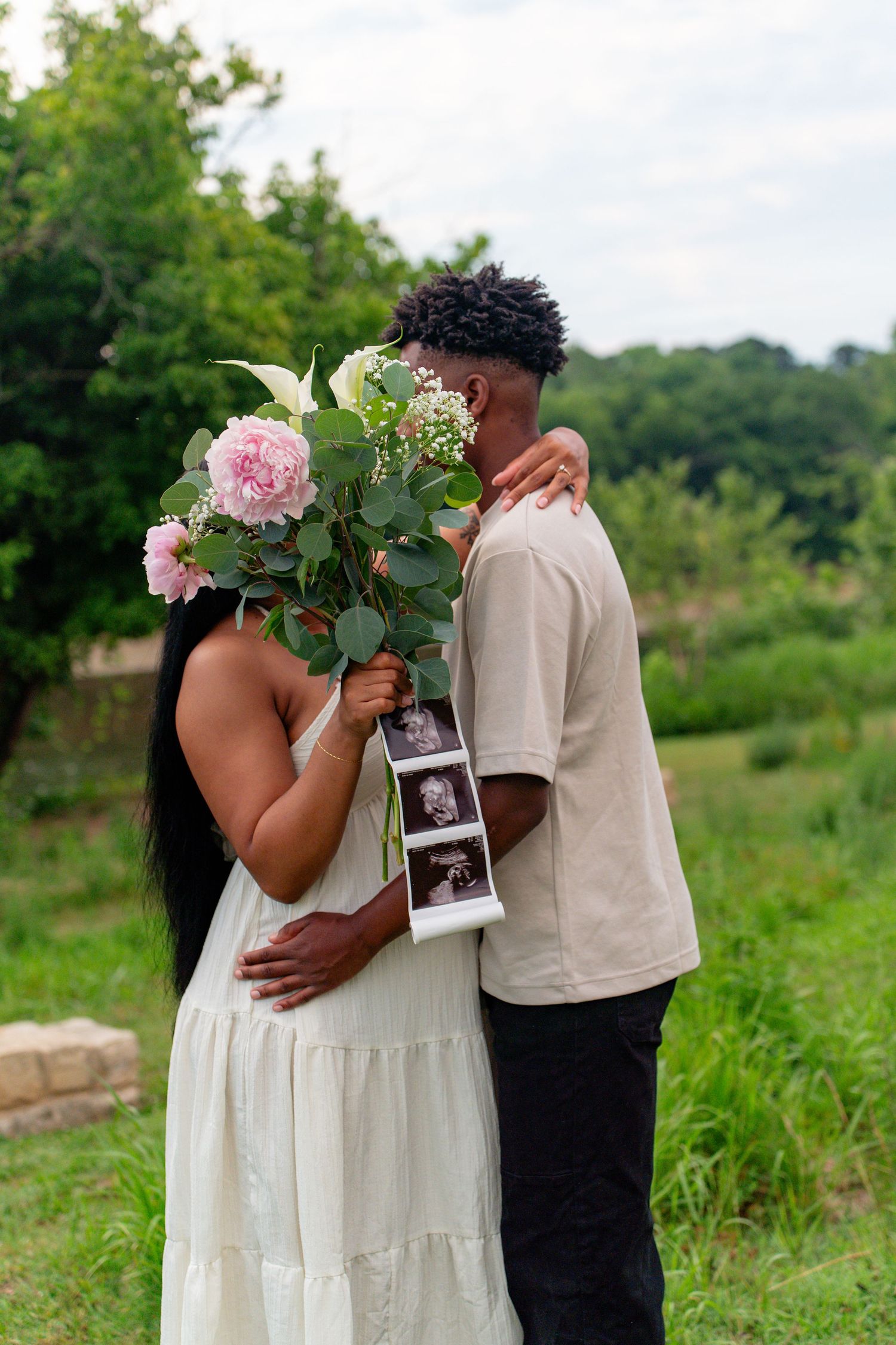 Couple holding pink flowers and ultrasound photos outdoors, pregnancy announcement in a lush green garden setting.