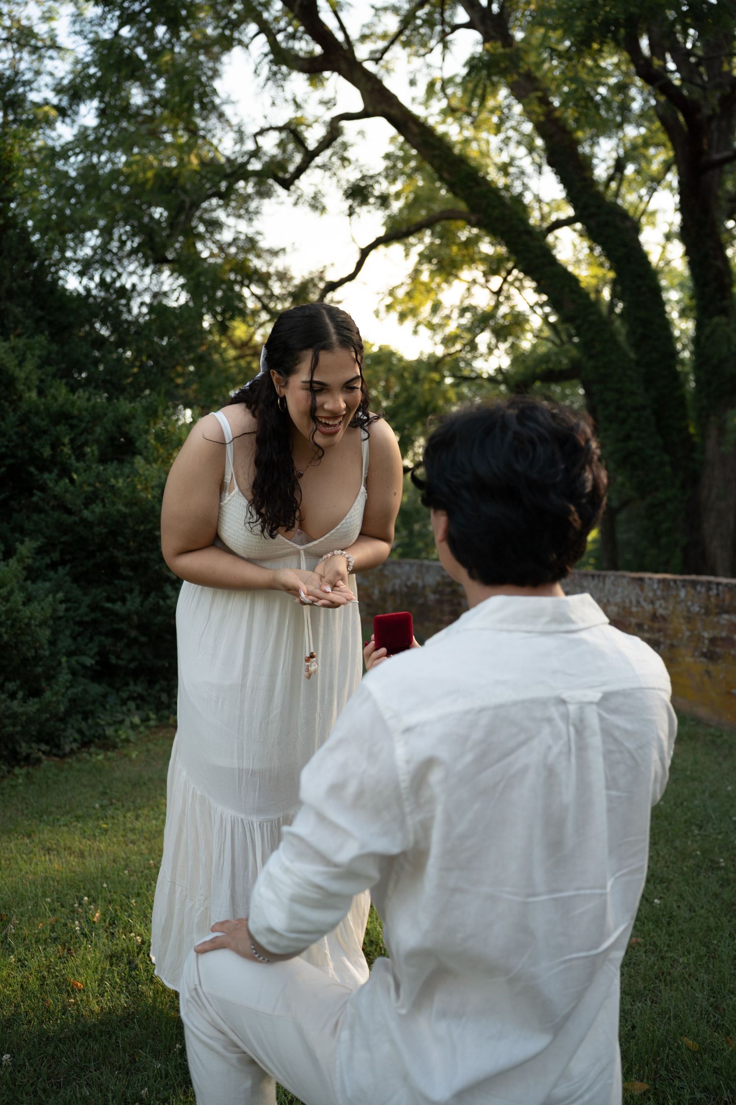 Man kneeling on one knee proposing to joyful woman in white dress outdoors at dusk. Taken by Row And Co Photography in FXBG VA