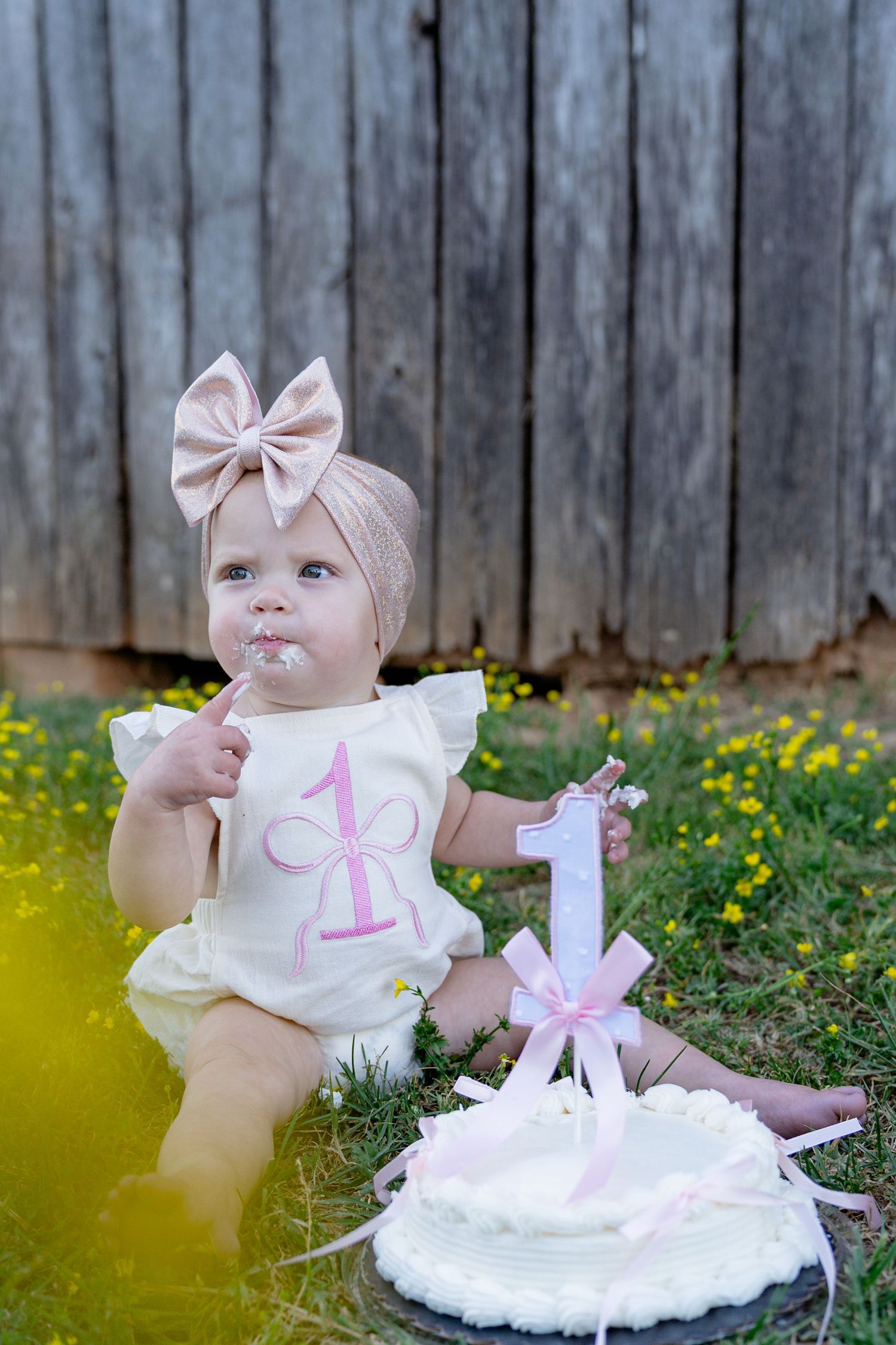 A baby girl in a white romper celebrates her first birthday with a smash cake outdoors. Taken by Row And Co Photography in FXBG VA