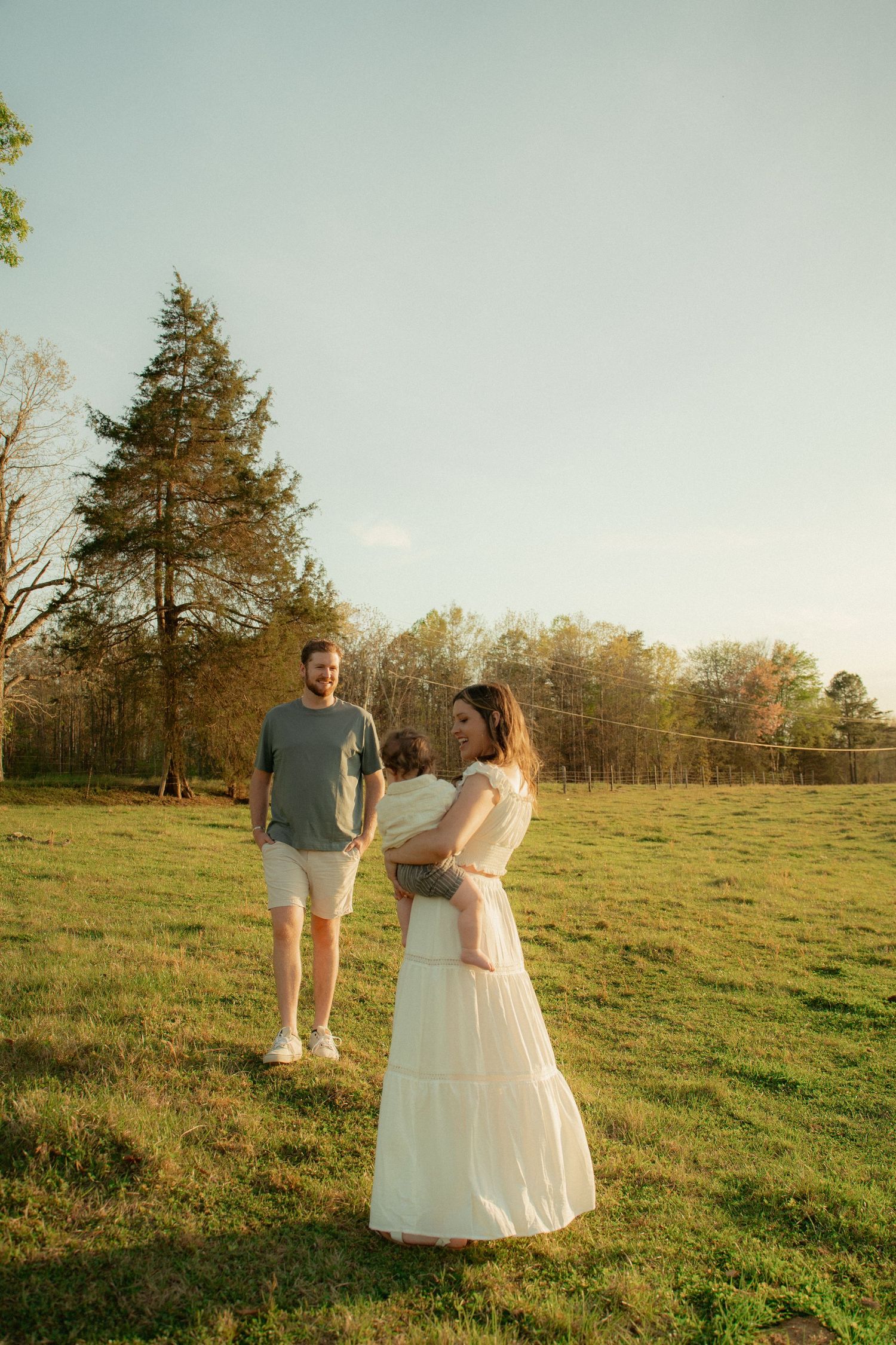 Family portrait of a woman in a white dress holding a baby while a man in a green shirt stands in a sunny green field. Taken by Row And Co