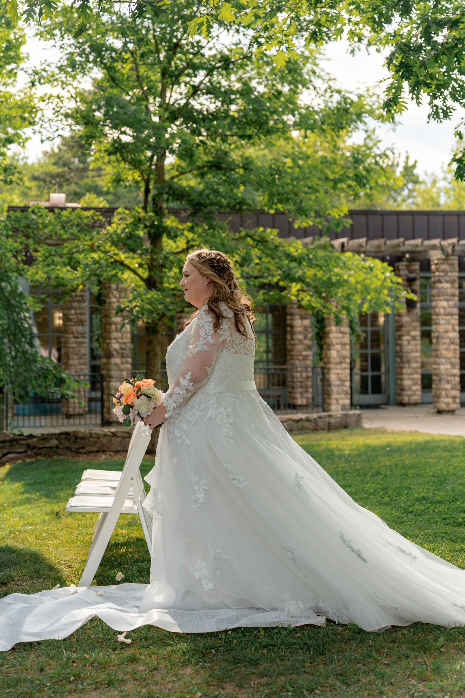 Bride in long-sleeve lace wedding gown holding bouquet beside white chair in lush garden with stone building backdrop. Row and Co Photograph